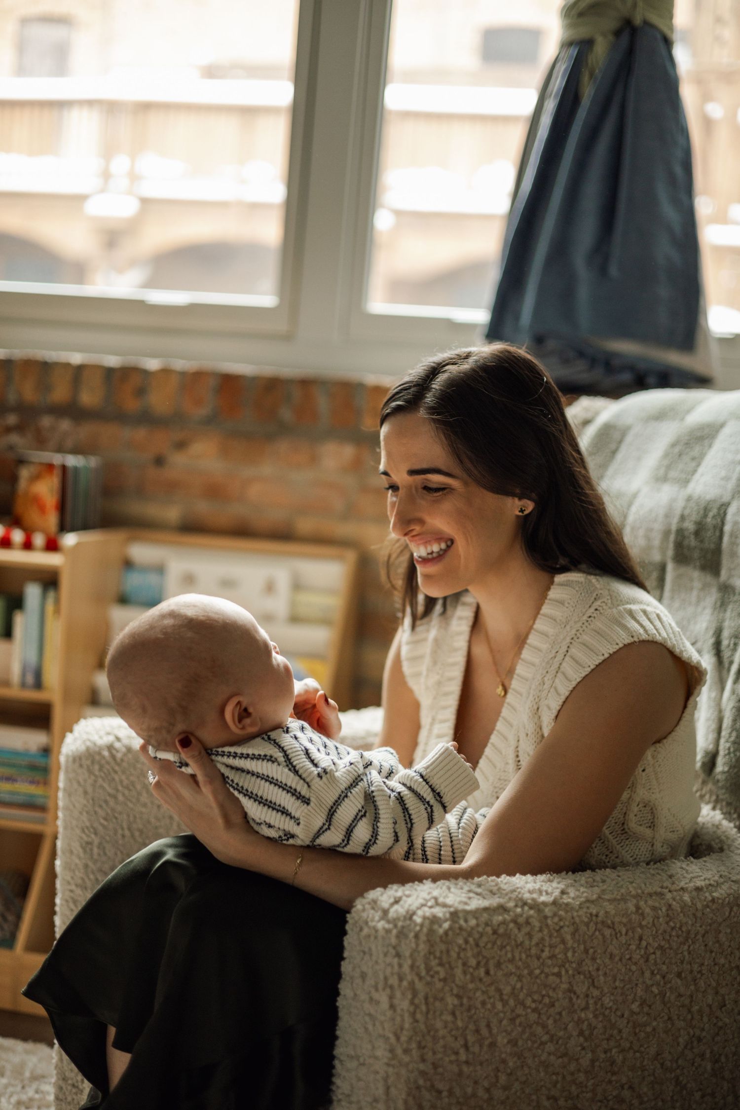 Smiling young mother sitting in armchair holding her newborn baby in a sunlit nursery room.