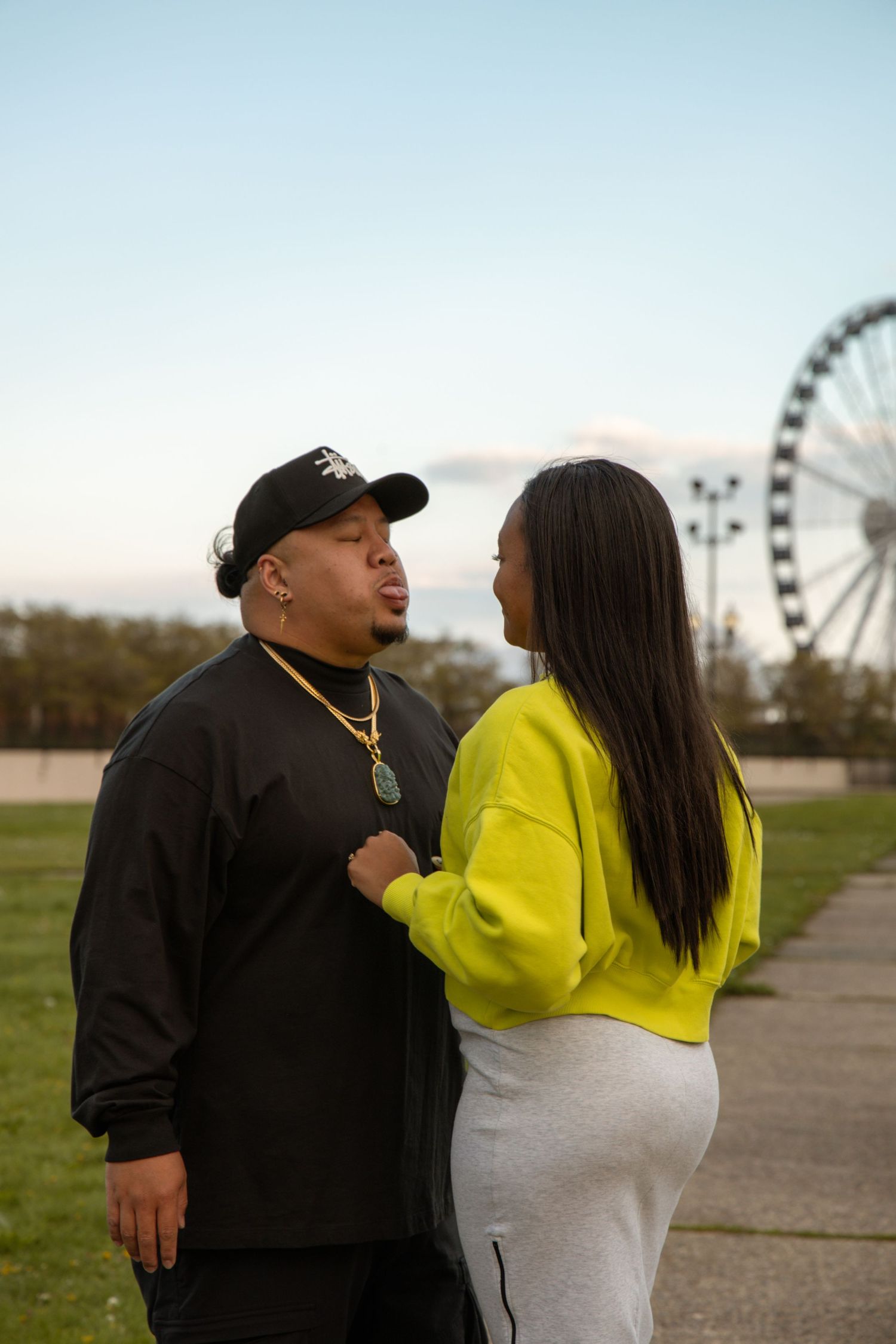 Couple shares romantic moment outdoors near Ferris wheel at dusk, woman in yellow jacket faces man in black.