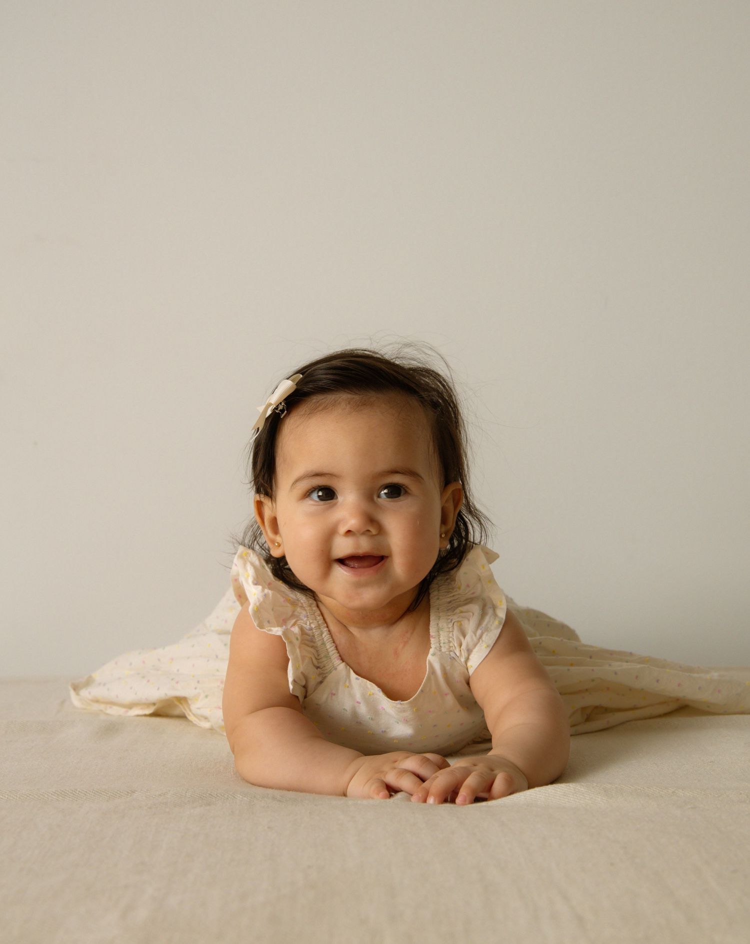 Smiling baby girl in cream lace dress doing tummy time on beige fabric backdrop.