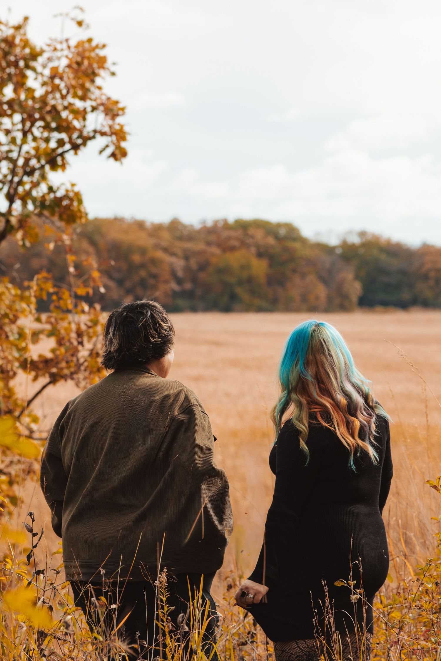 Couple standing back-to-back in autumn field, woman with blue hair, overlooking golden landscape.