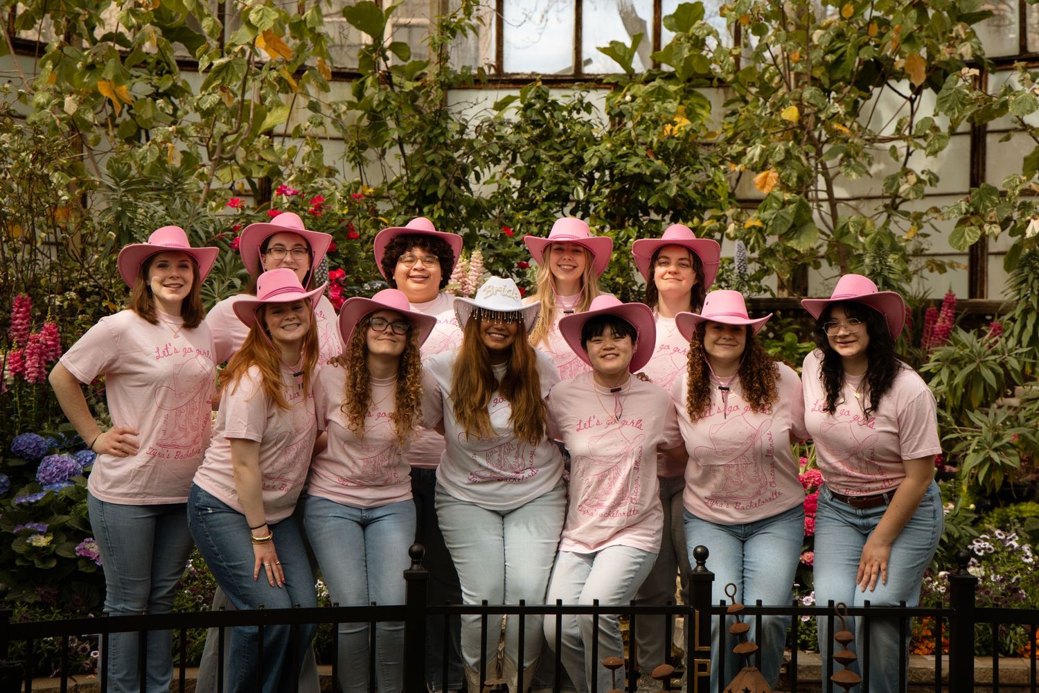 Group of women in matching pink t-shirts and cowboy hats posing outdoors among lush green plants.