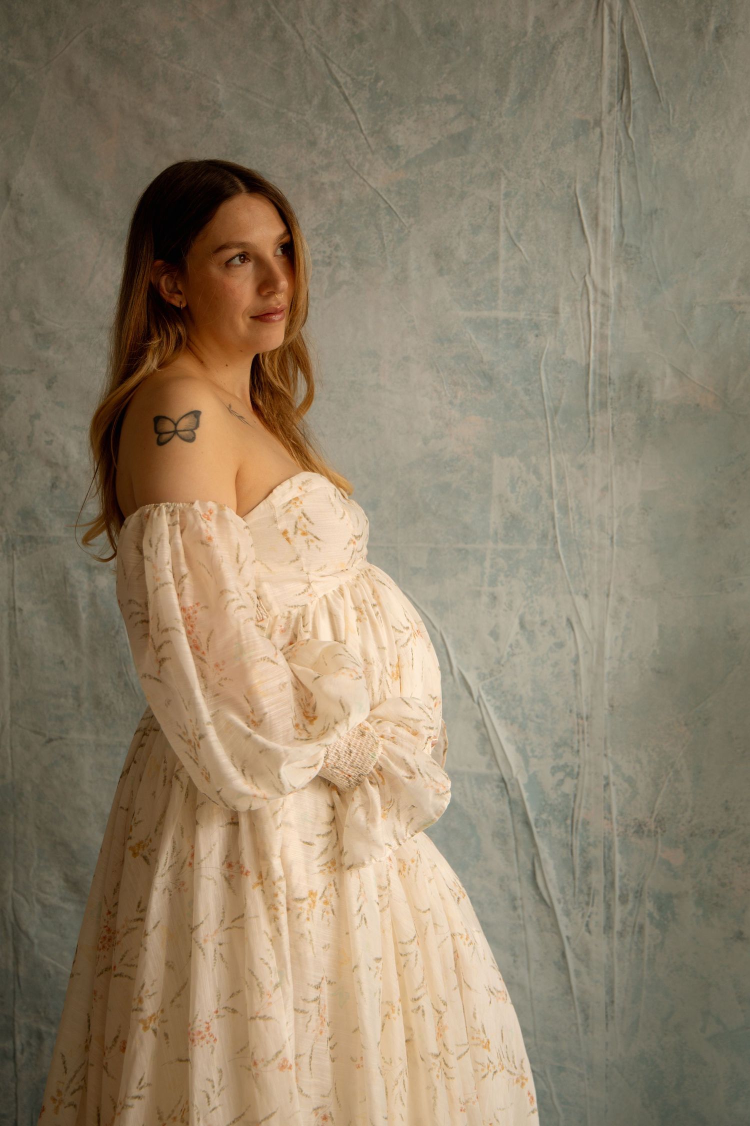 Pregnant woman in off-shoulder floral dress holding belly, butterfly tattoo, posing against gray backdrop.