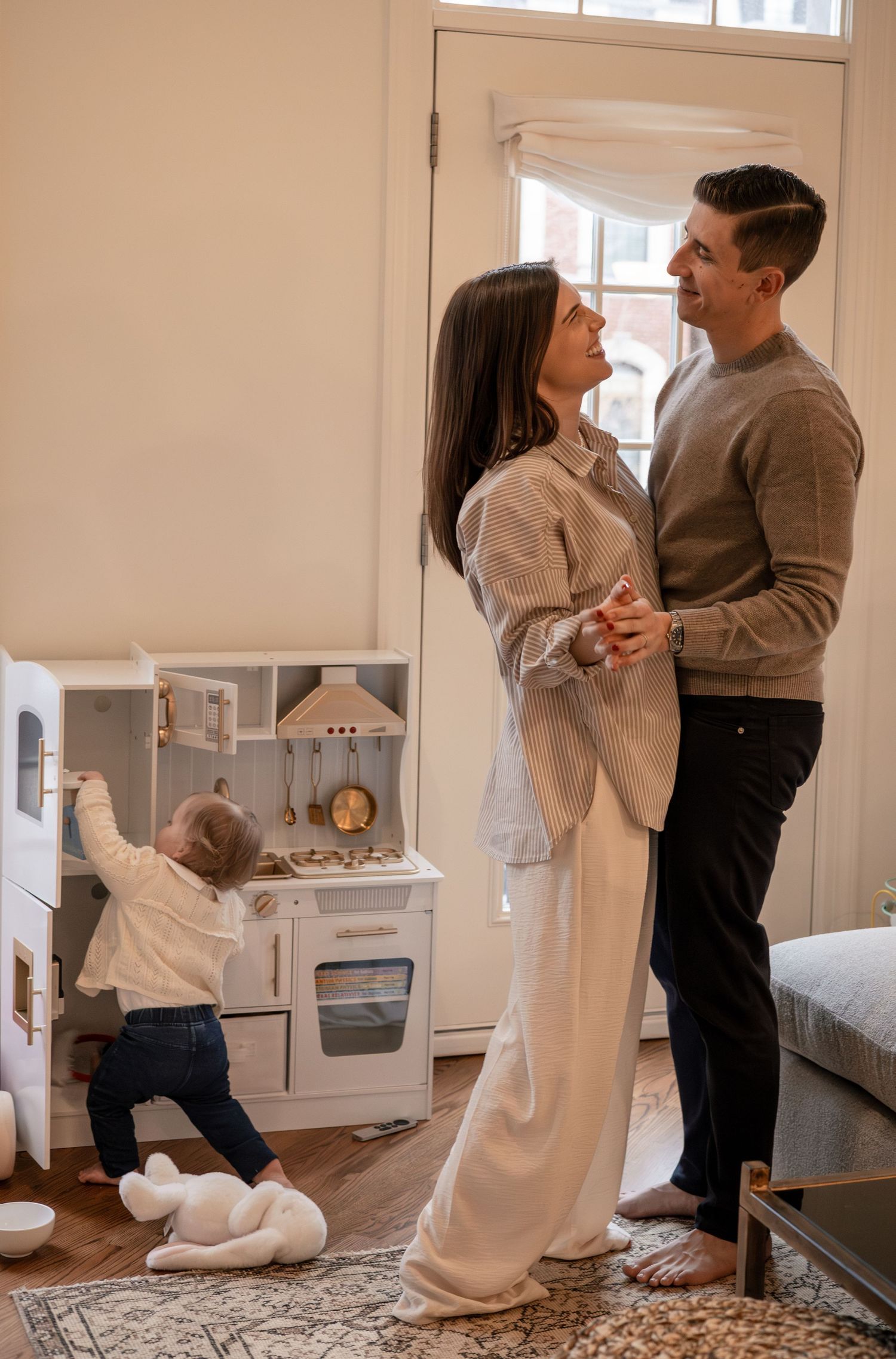 A couple shares a tender moment while their toddler plays with a white toy kitchen in a cozy living room.