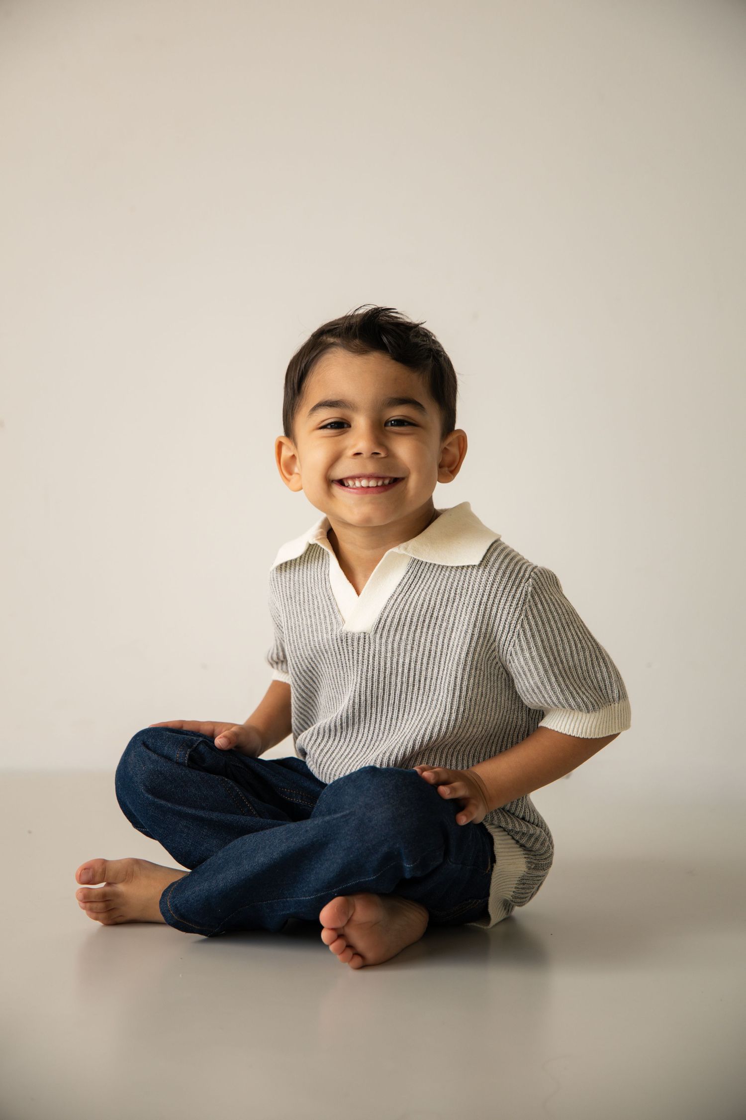 Smiling young boy sitting cross-legged on floor wearing striped polo shirt and jeans in studio photo.