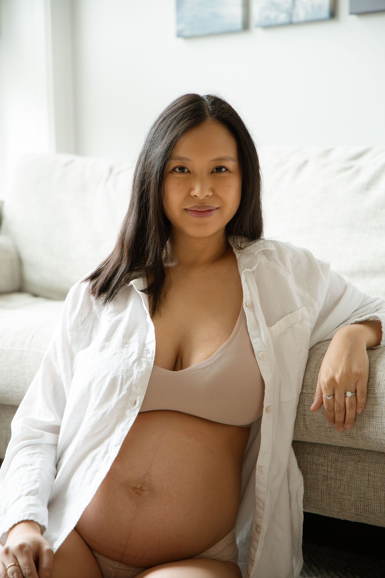 Smiling pregnant Asian woman in white shirt and nude bra sitting against couch, showing baby bump.