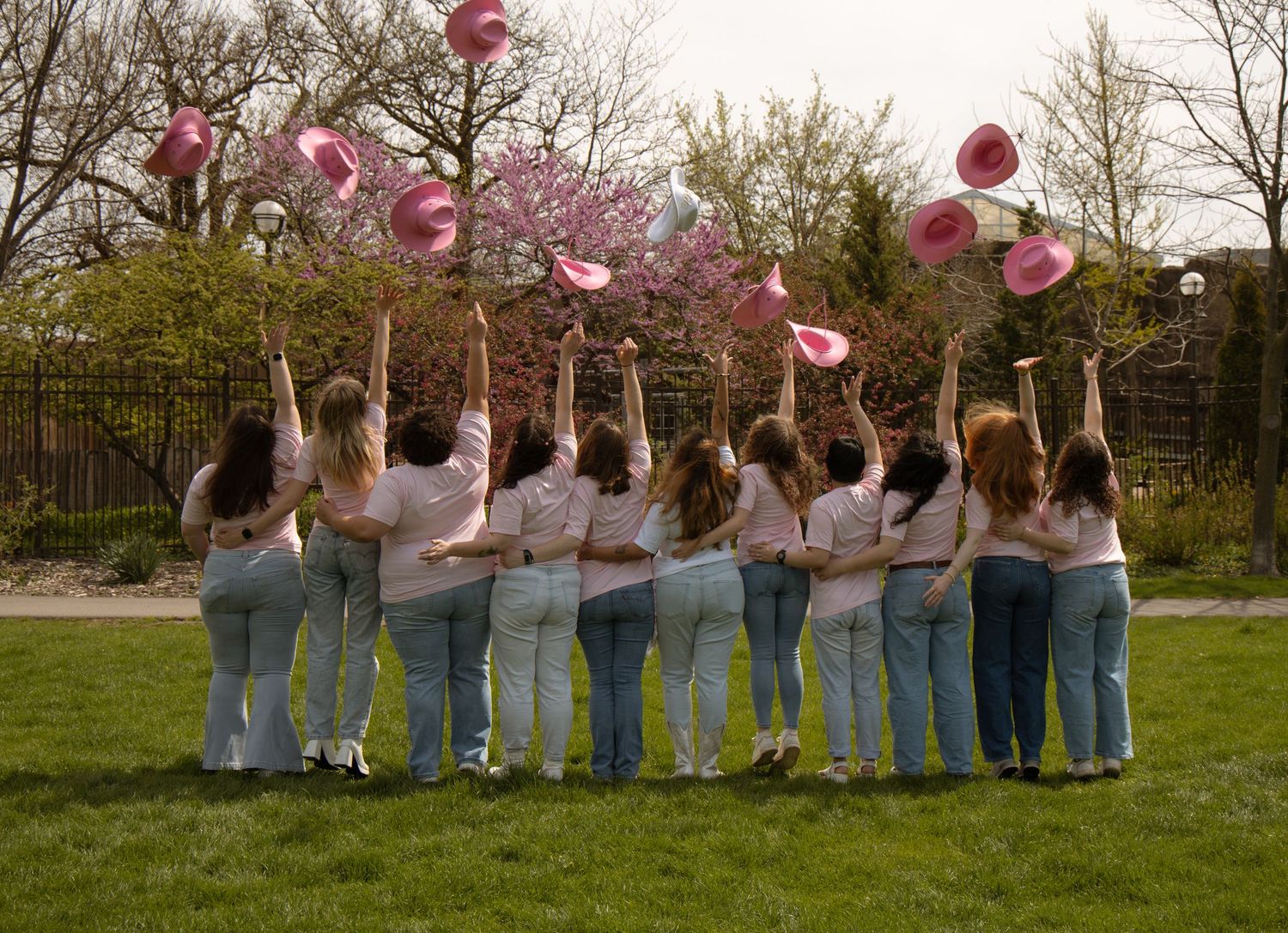 Group of women in white shirts and jeans tossing pink balloons and cowboy hats in a park.