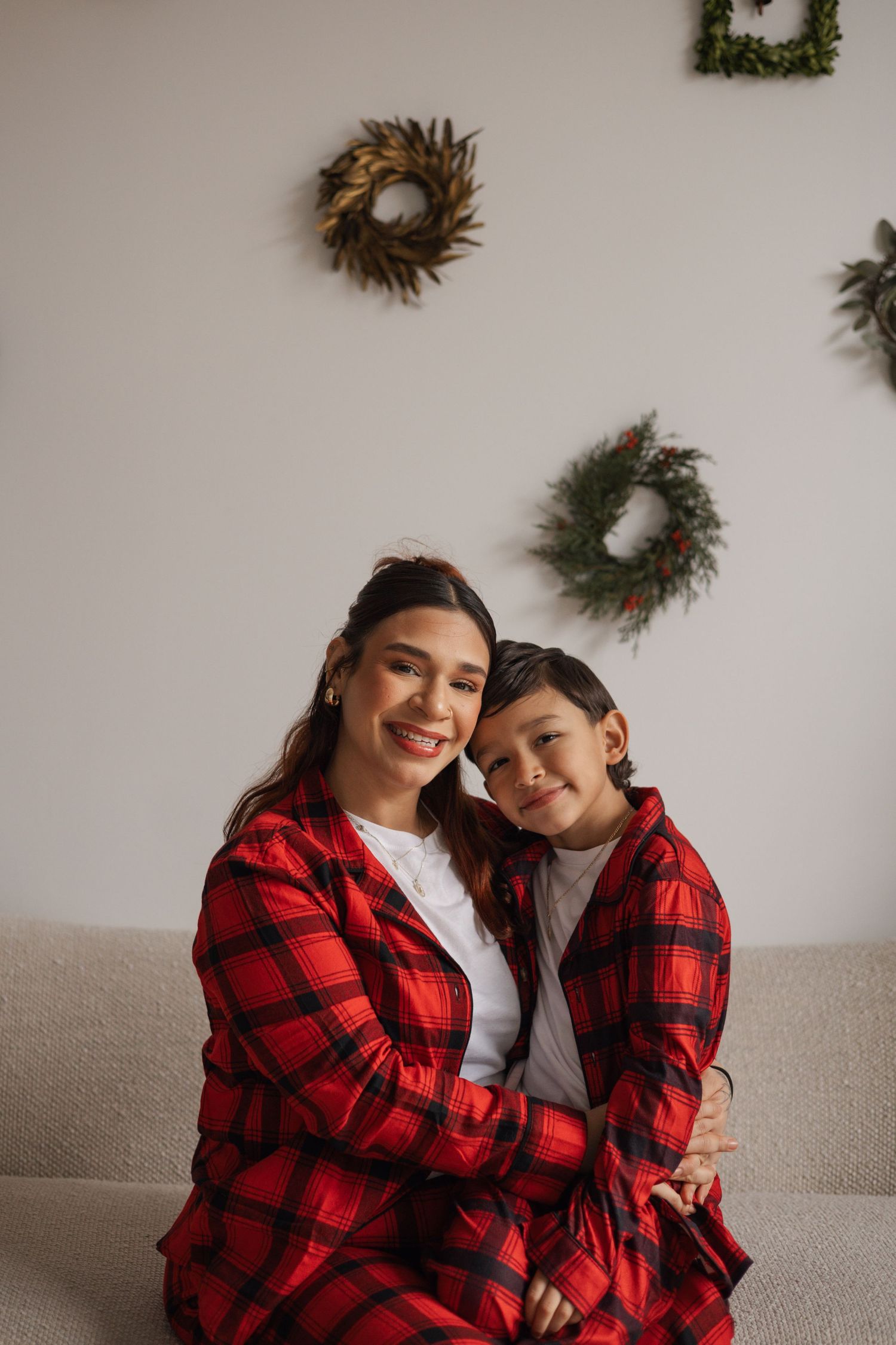 Mother and child in matching red plaid outfits embrace warmly on a sofa with holiday wreaths behind.