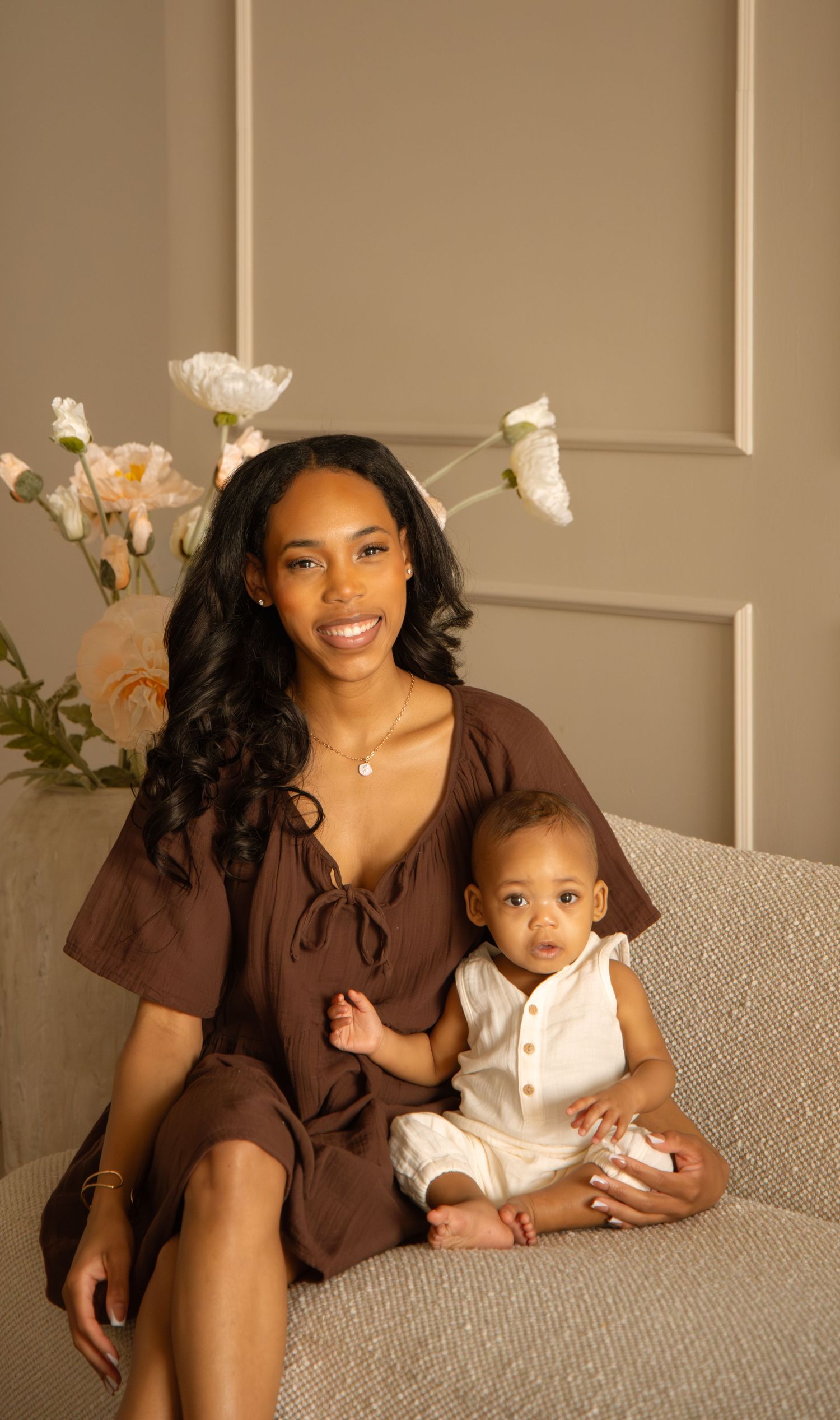 A young woman in a brown dress sits with her baby in a white romper, posed in front of white flowers.
