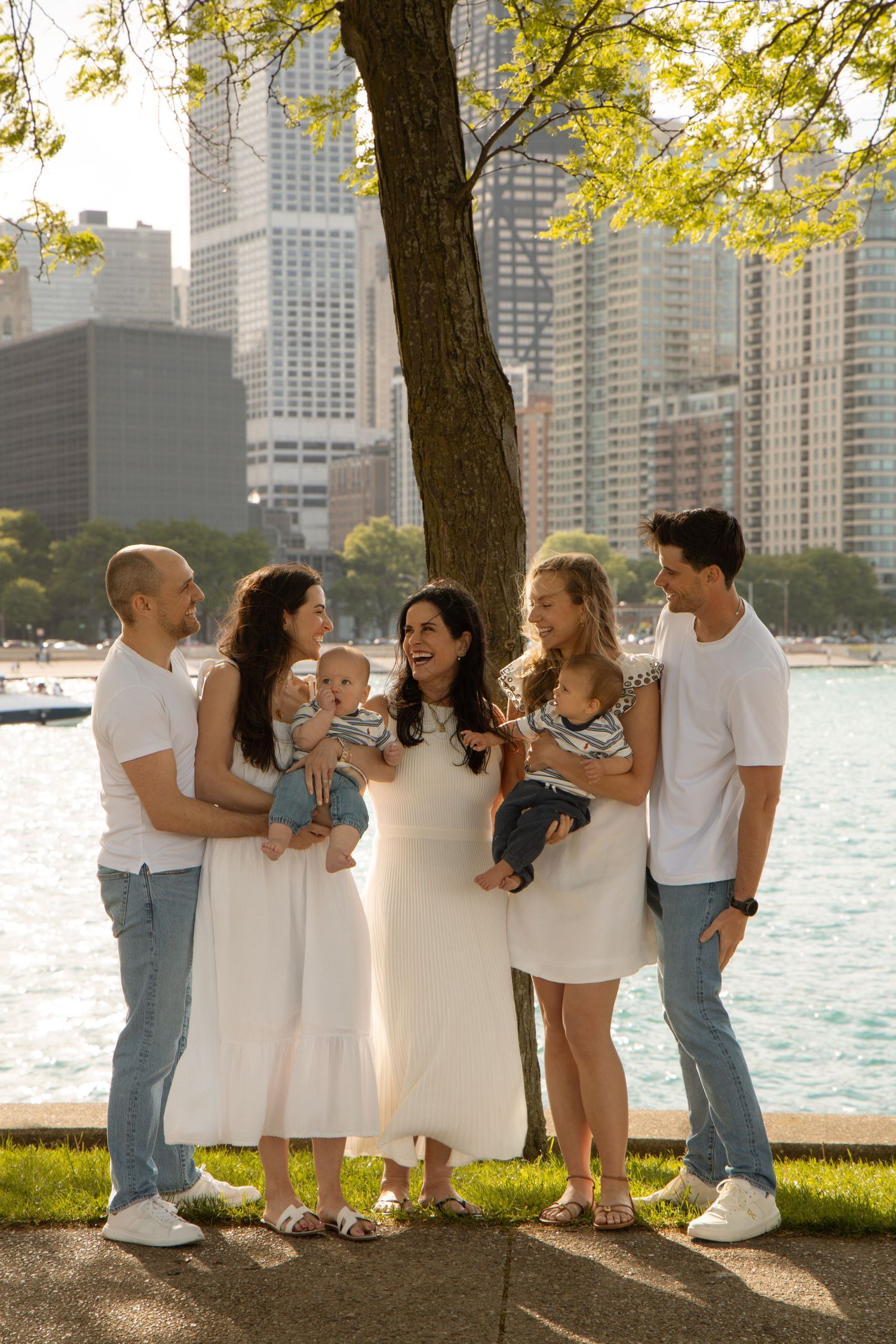 A joyful family of six poses near a city waterfront, two couples holding babies, surrounded by lush trees and skyline.