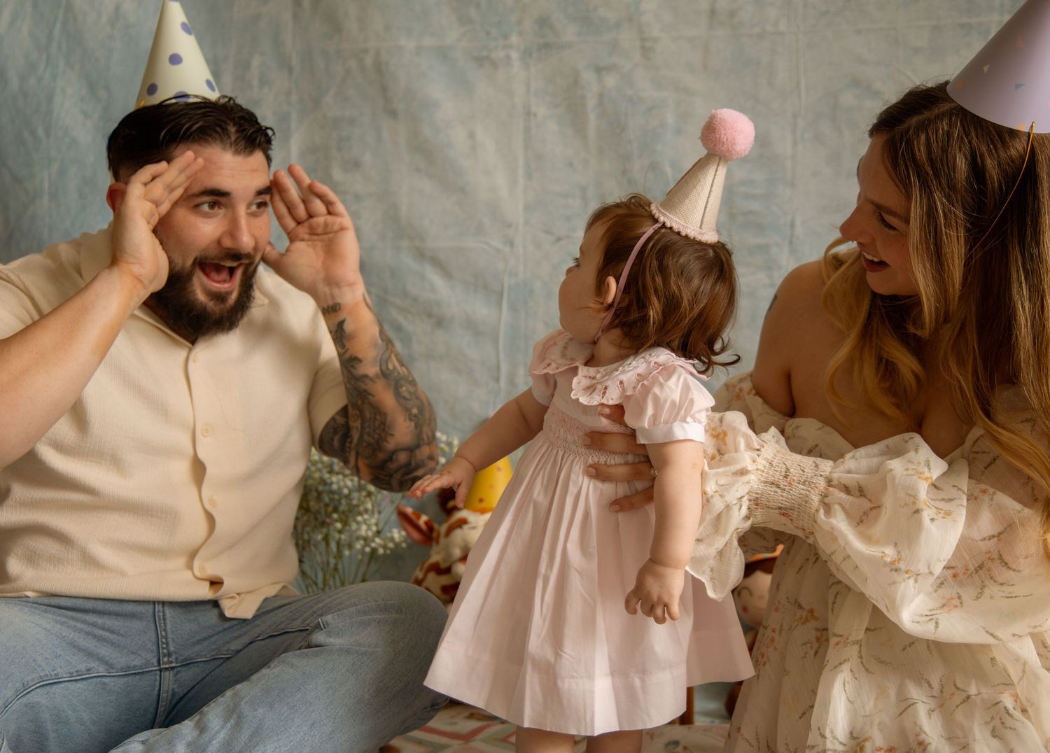 Family celebrating toddler birthday party with party hats, parents laughing joyfully with baby girl in white dress.