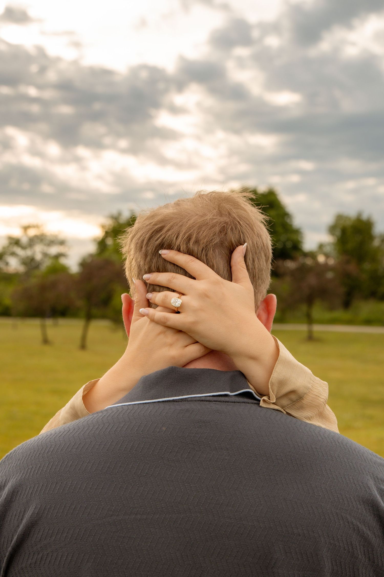 A woman's hand with a gold ring rests on the back of a man's head in a park setting.