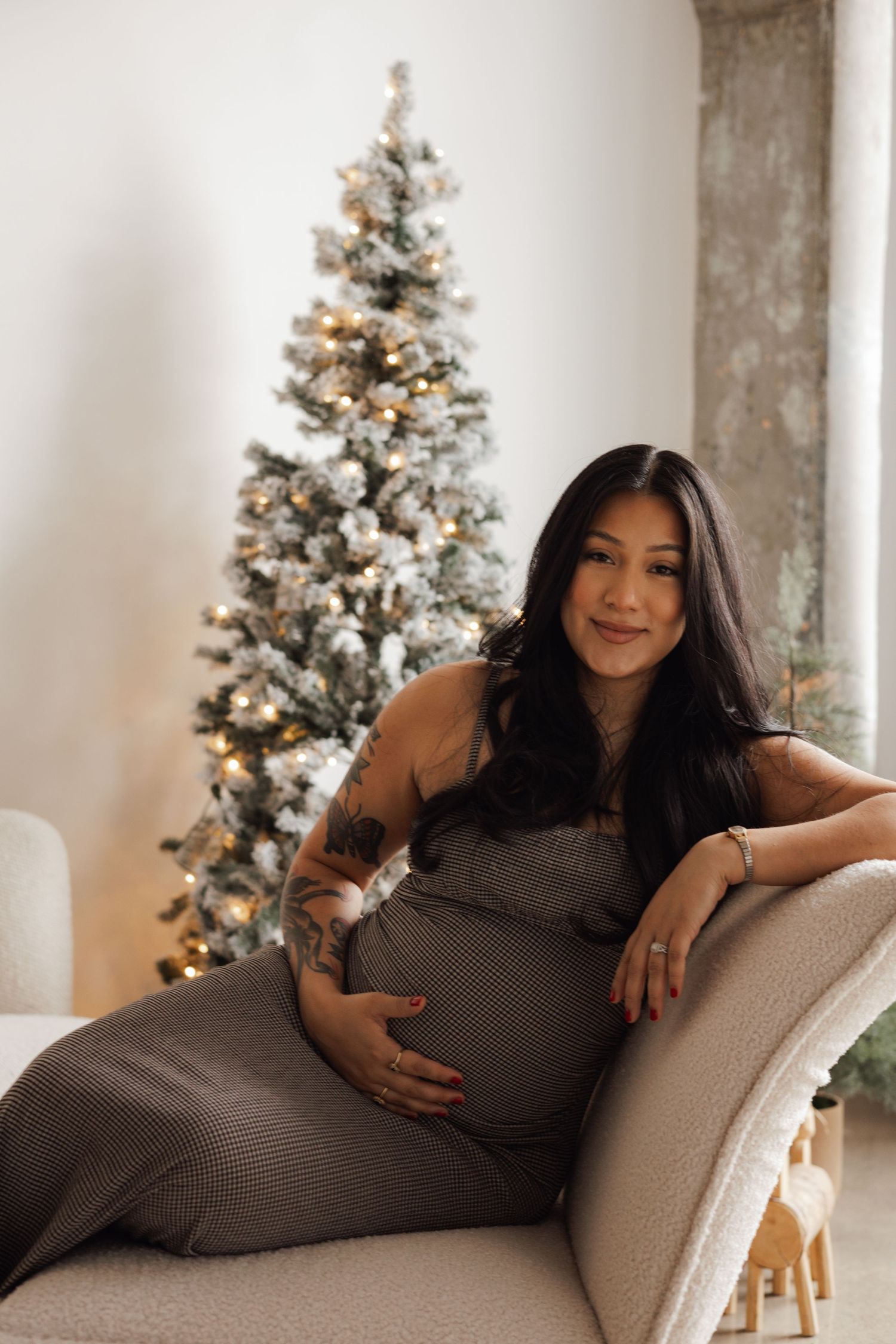 Pregnant woman in gray dress sitting on a chair near a decorated Christmas tree indoors.