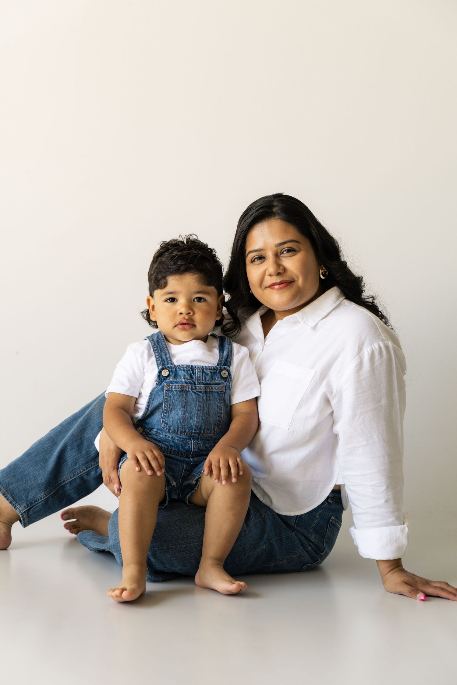 A woman and toddler in denim overalls sit together smiling against a white background.
