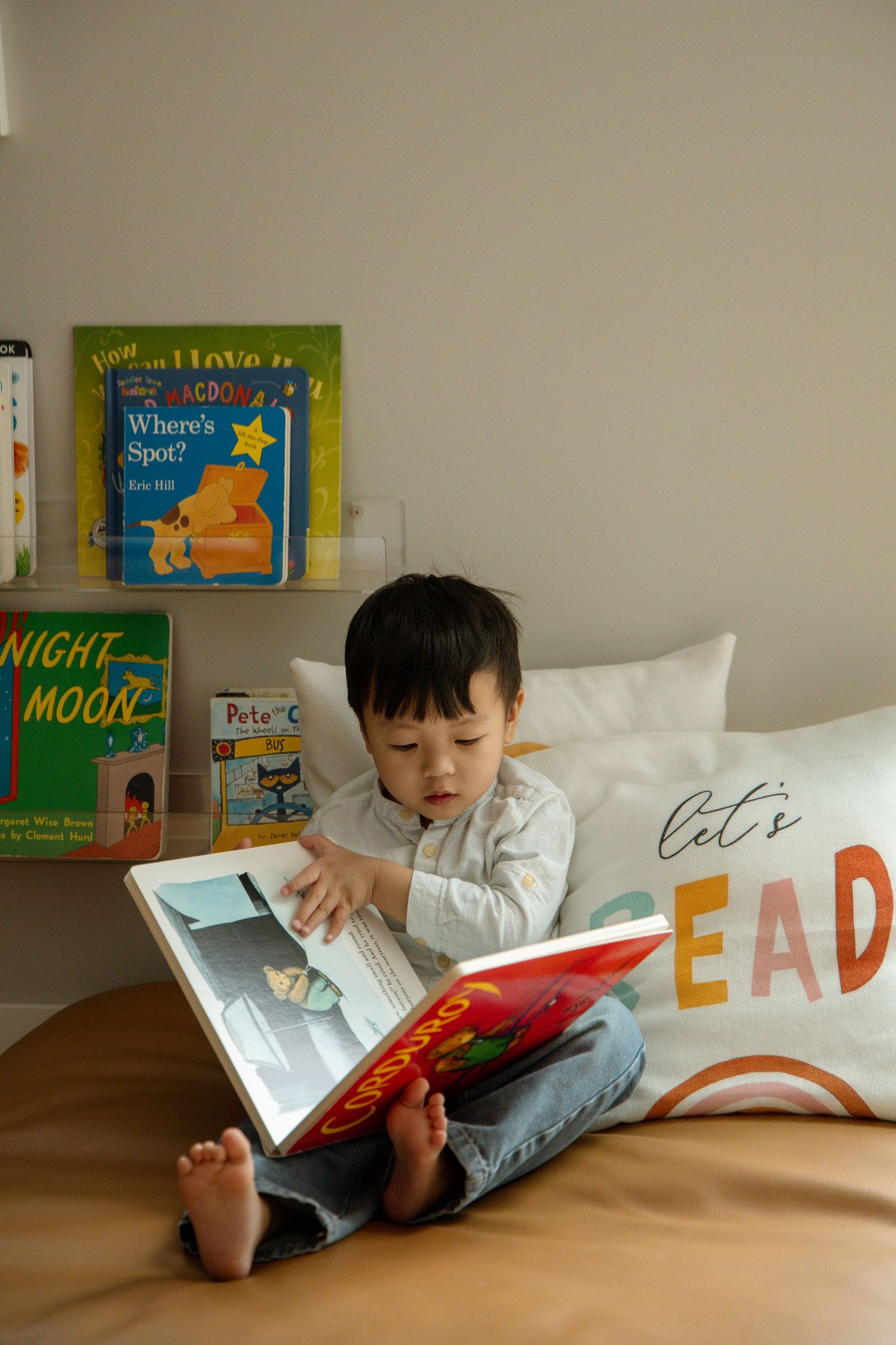 Young boy sitting cross-legged on bed reading a colorful book, with children's books on shelf behind him.