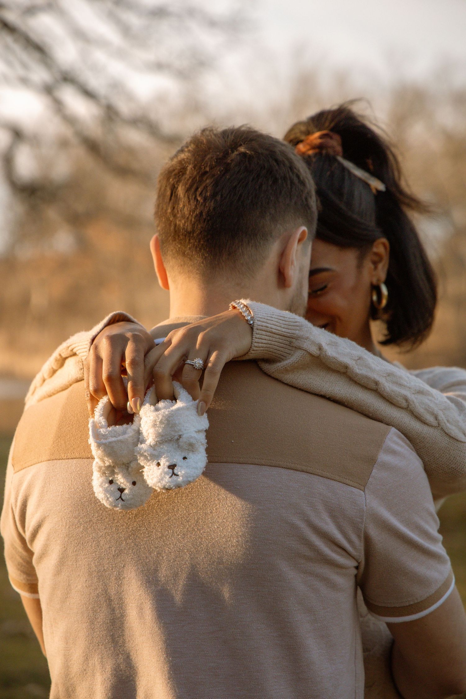 Couple embracing outdoors at sunset, woman holding tiny white baby booties over mans shoulder.