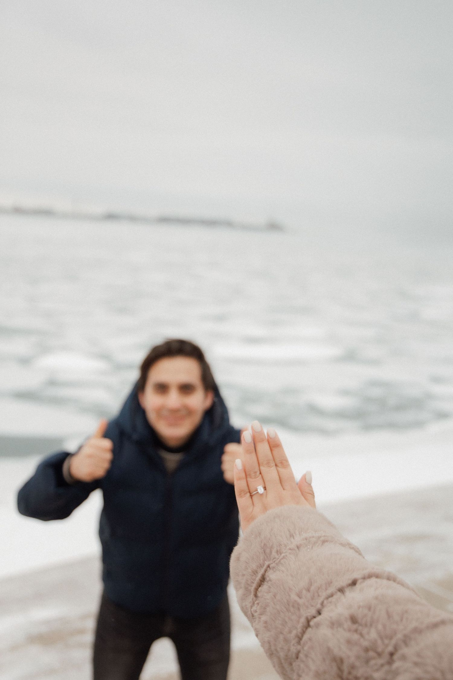 Excited man giving thumbs up as woman displays engagement ring near frozen Lake Michigan shoreline.