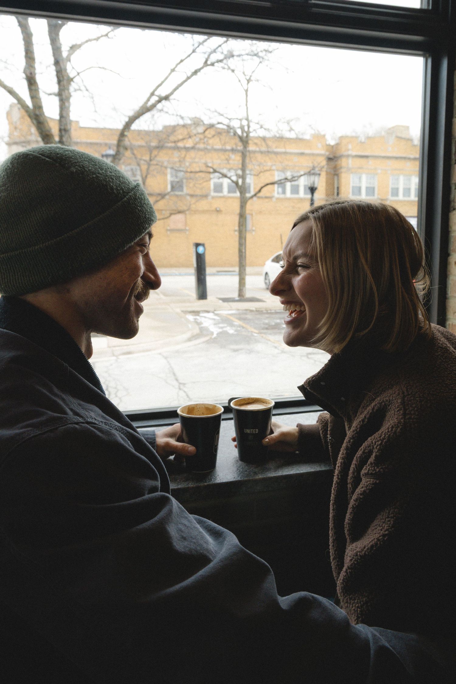 Couple laughing together at coffee shop window seat, each holding espresso cups on winter day.