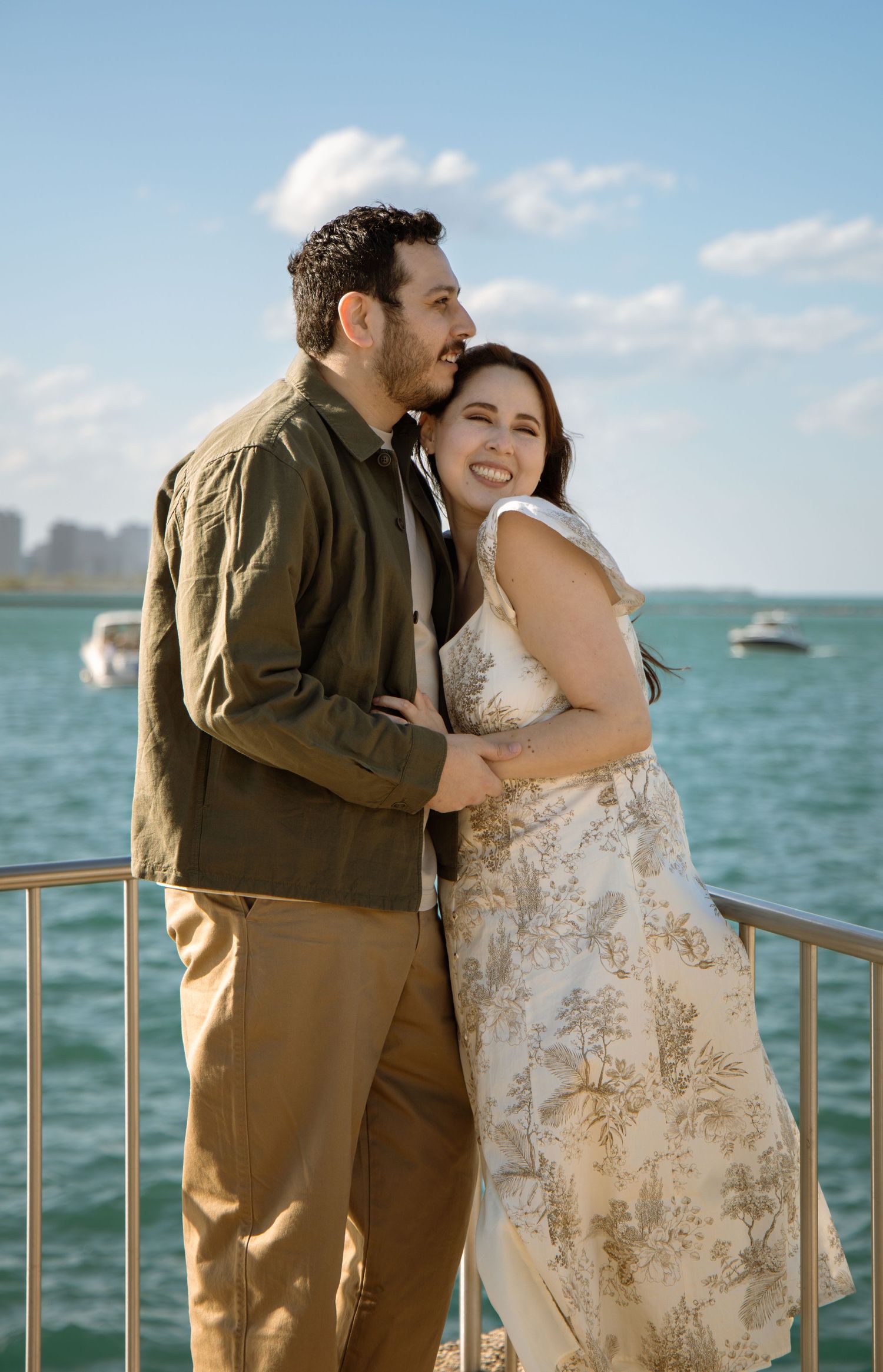 A smiling couple embraces on a waterfront pier, woman in floral dress and man in olive shirt, with blue water behind them.