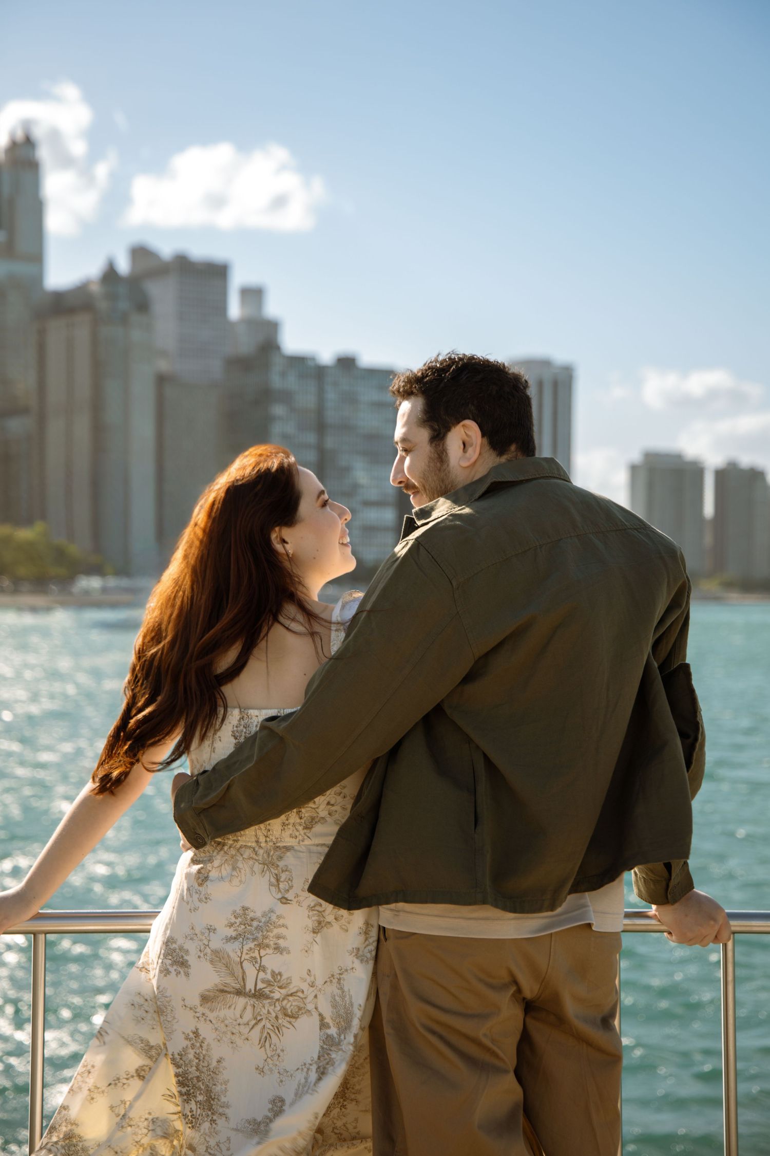 Couple embracing by waterfront railing with city skyline backdrop on a sunny day.