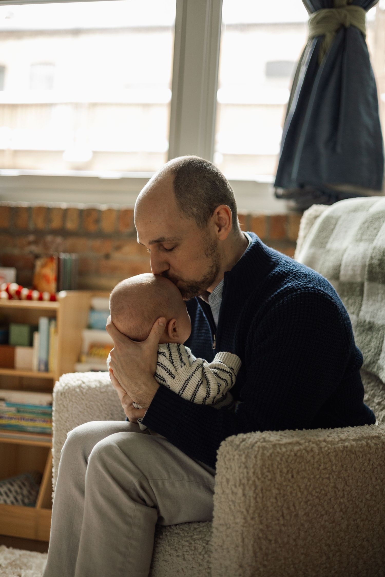A bald father tenderly kisses his newborn baby while seated in a cozy armchair near a bright window.