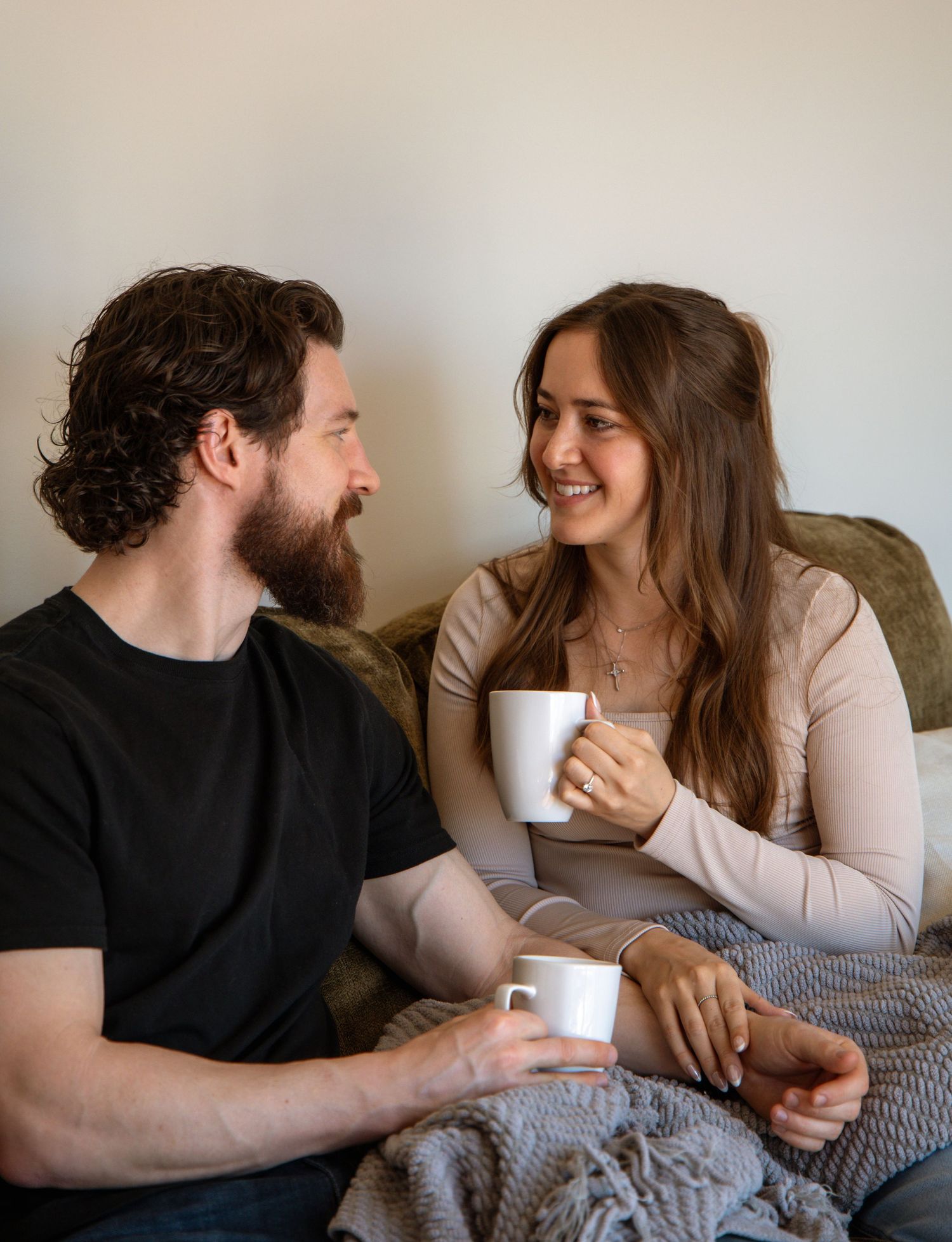 A smiling couple sits on a couch sharing coffee mugs in a bright, relaxed home setting.