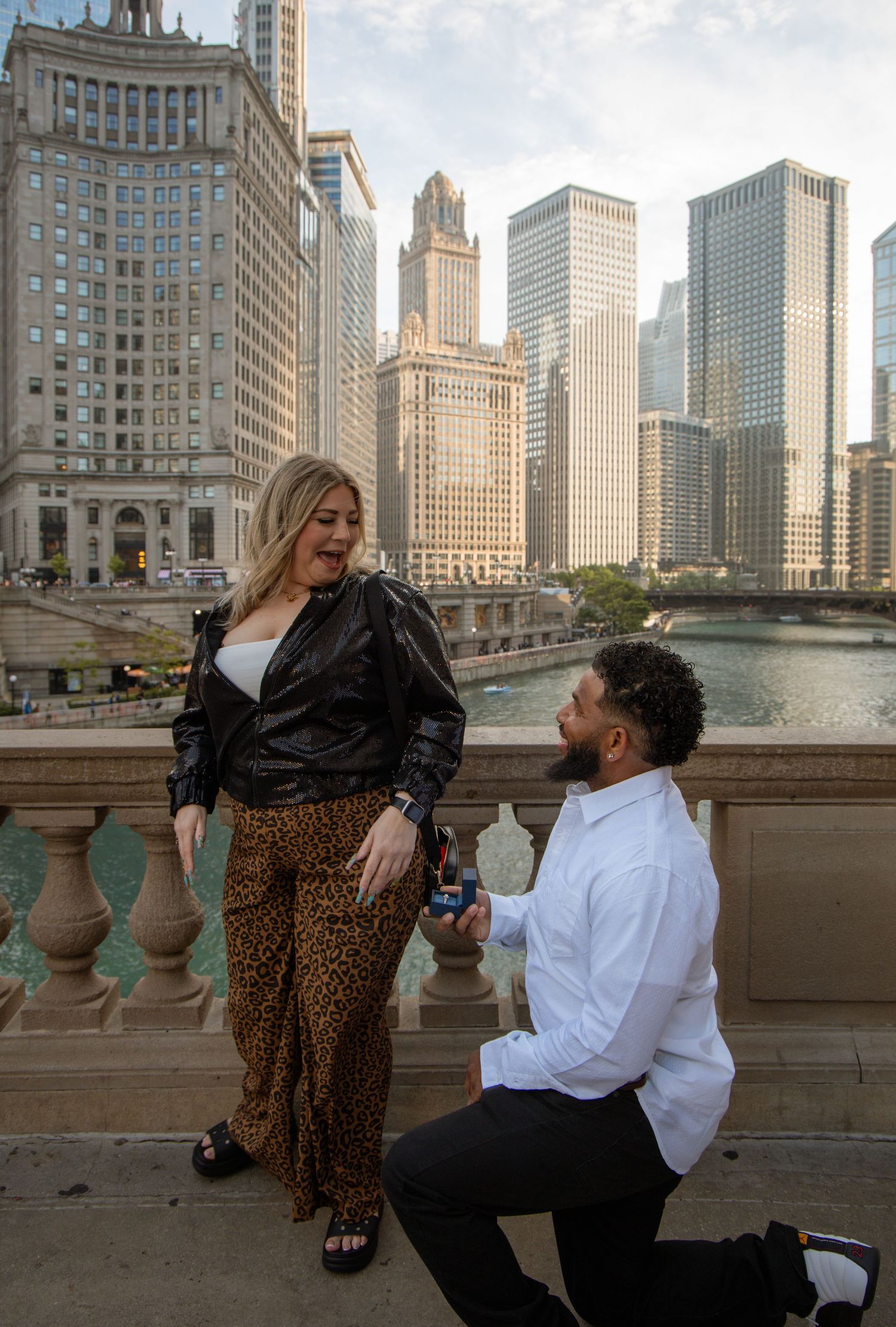 Man proposes on one knee to woman on Chicago Riverwalk with city skyline in background.