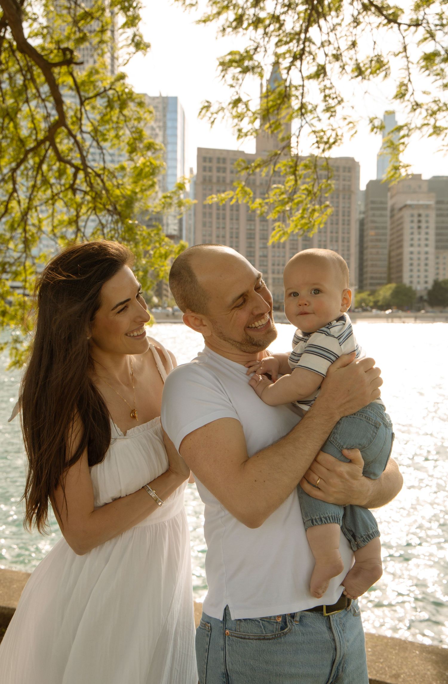 A couple smiles at their baby boy by a city waterfront on a sunny day, woman in white dress, man in white tee.