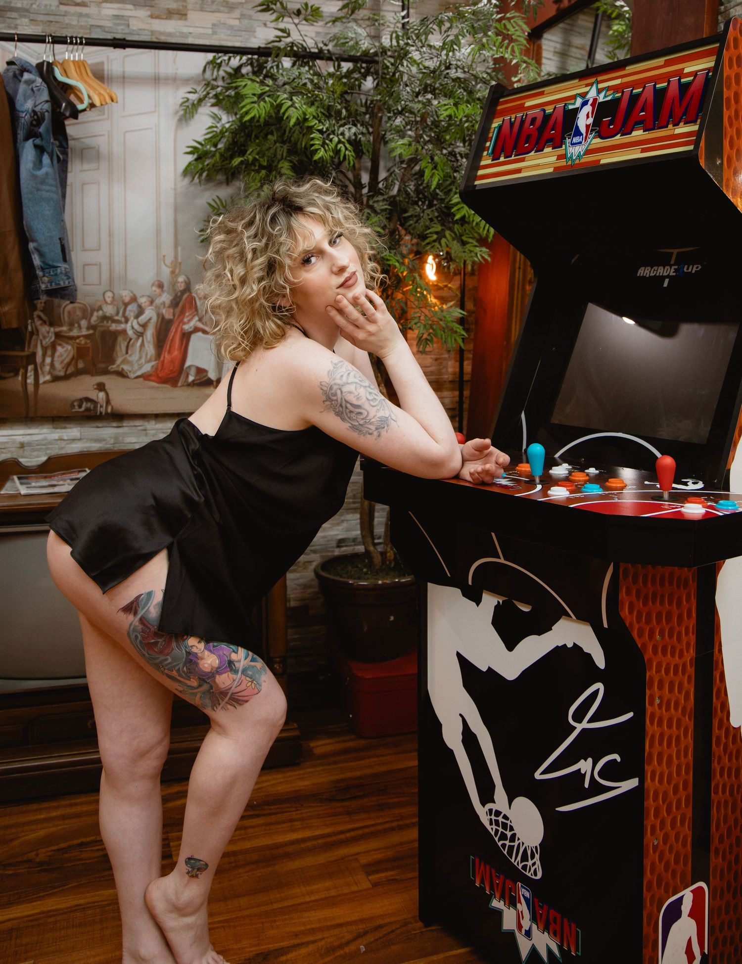 Tattooed woman in black dress posing next to NBA Jam arcade game.