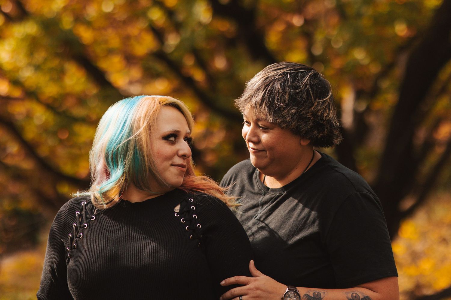 Two women smile at each other outdoors surrounded by vibrant golden autumn foliage.