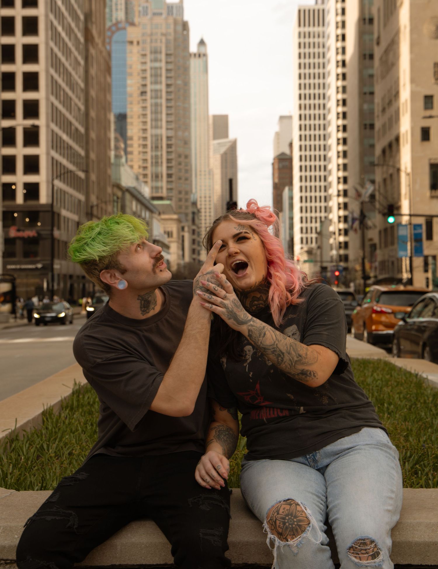Couple with colorful hair and tattoos laughing together on a city street bench surrounded by tall buildings.