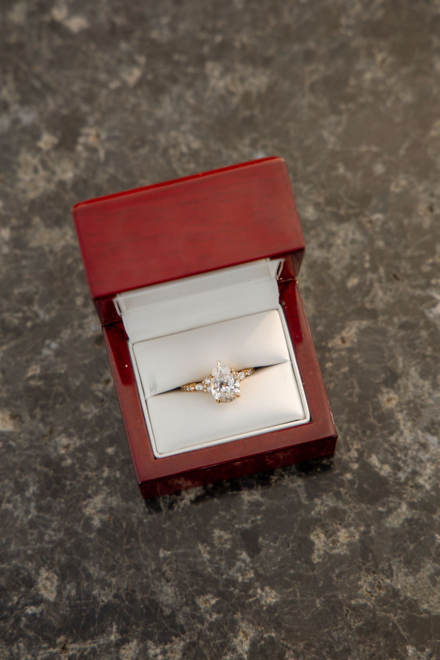 Gold solitaire diamond engagement ring displayed in an open red jewelry box on a marble surface.