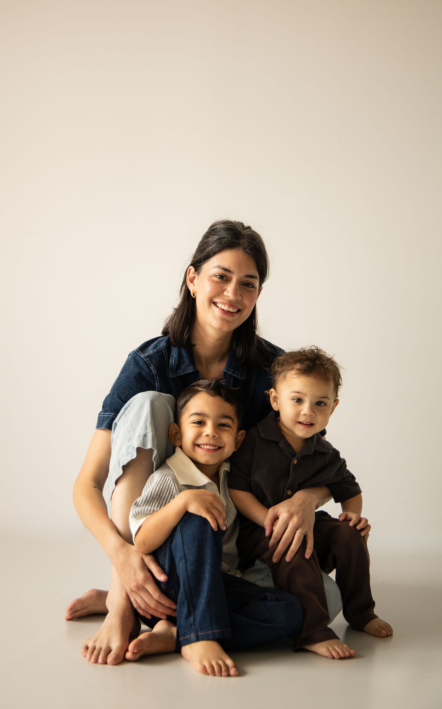 A smiling woman sits on the floor with two young children in a bright studio portrait.
