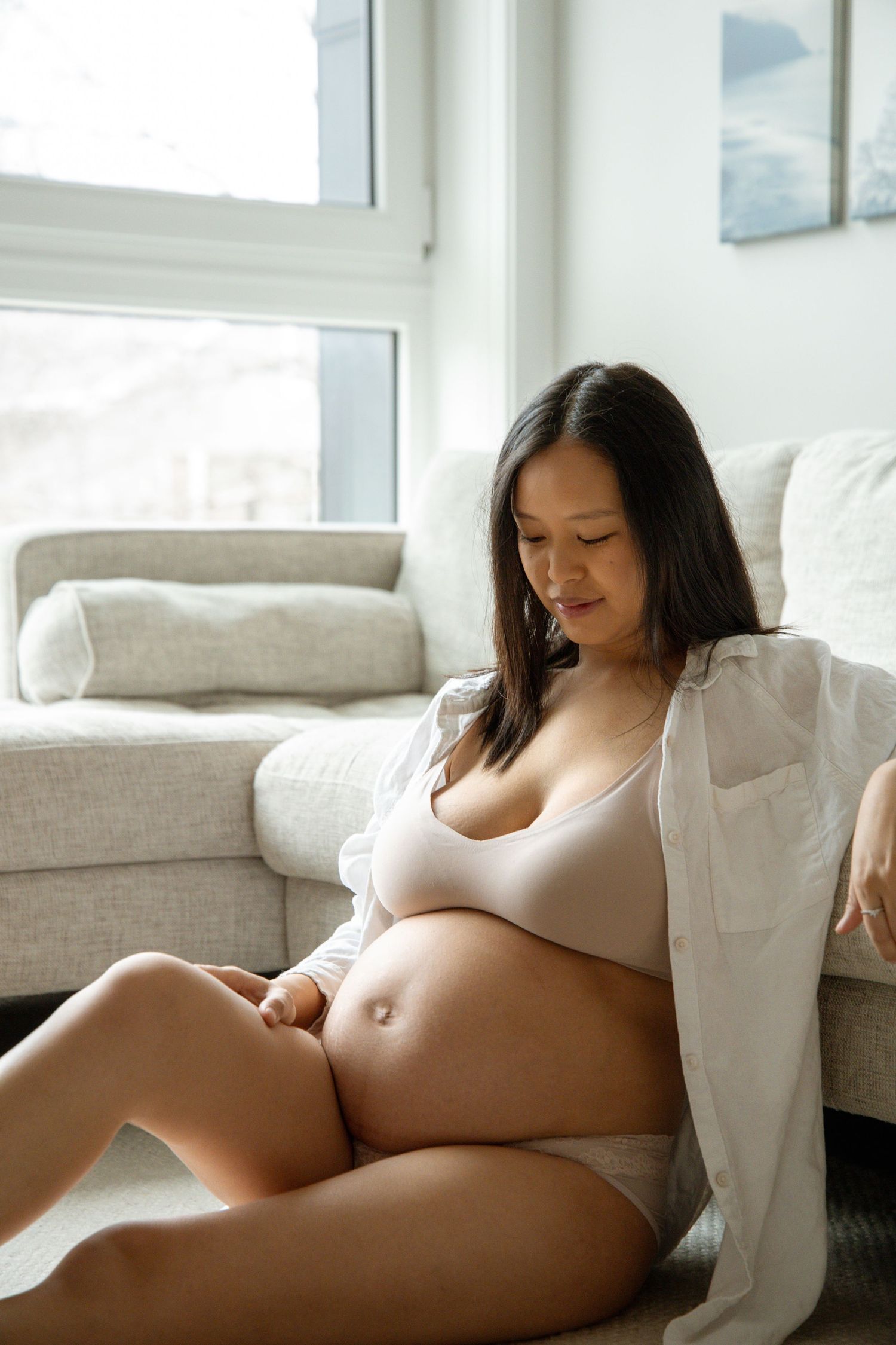 Pregnant Asian woman in beige lingerie and open white shirt sits on floor cradling her baby bump.