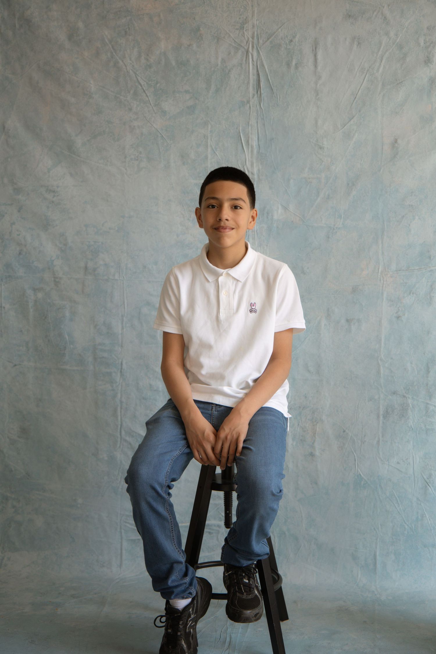 Young boy in white polo shirt and jeans sitting on a stool against a gray textured backdrop.