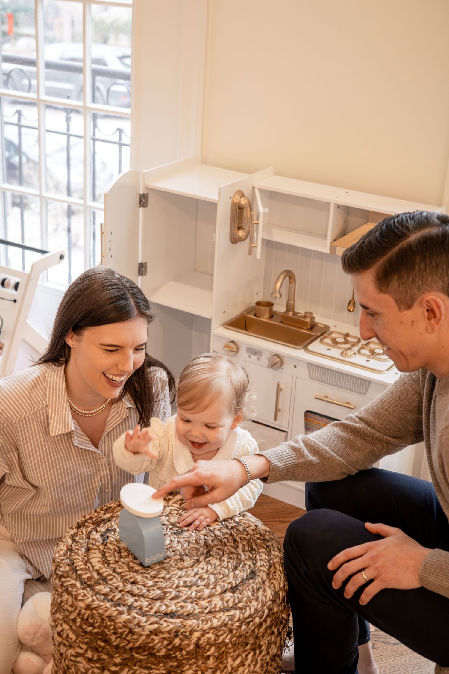 Happy family with laughing baby playing together near a toy kitchen set in a bright, cozy playroom.