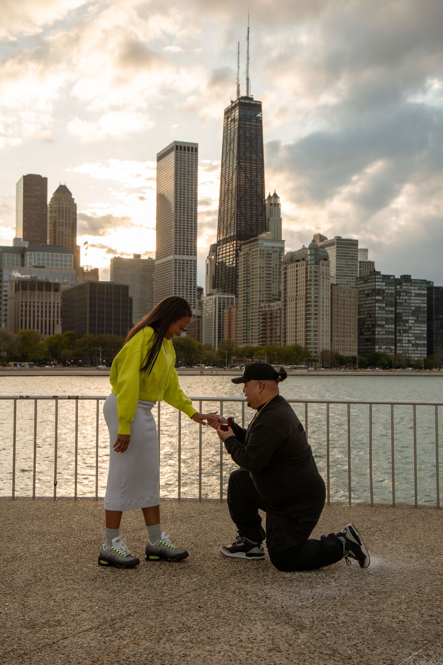 Man proposes on one knee to woman by Chicago waterfront with city skyline at sunset.