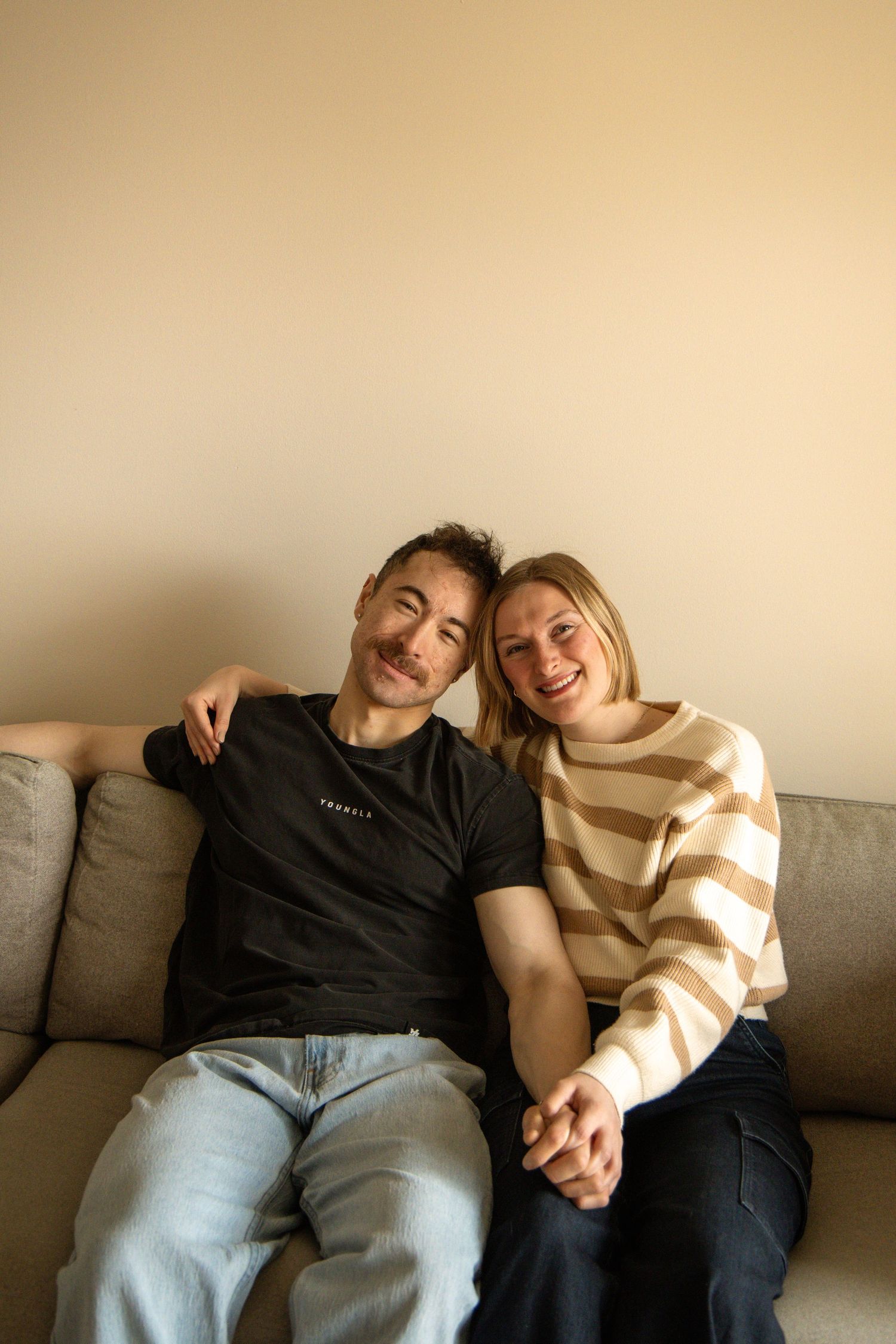 Smiling couple sitting on gray couch holding hands, man in black tee, woman in striped sweater.