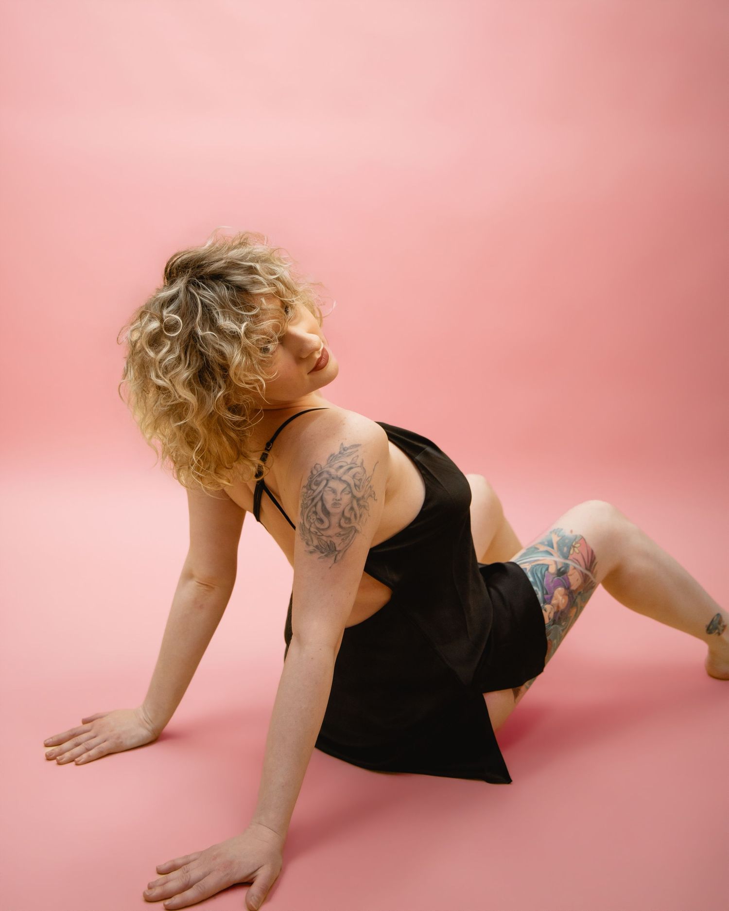 Tattooed blonde woman in black dress poses on pink studio floor with curly hair.