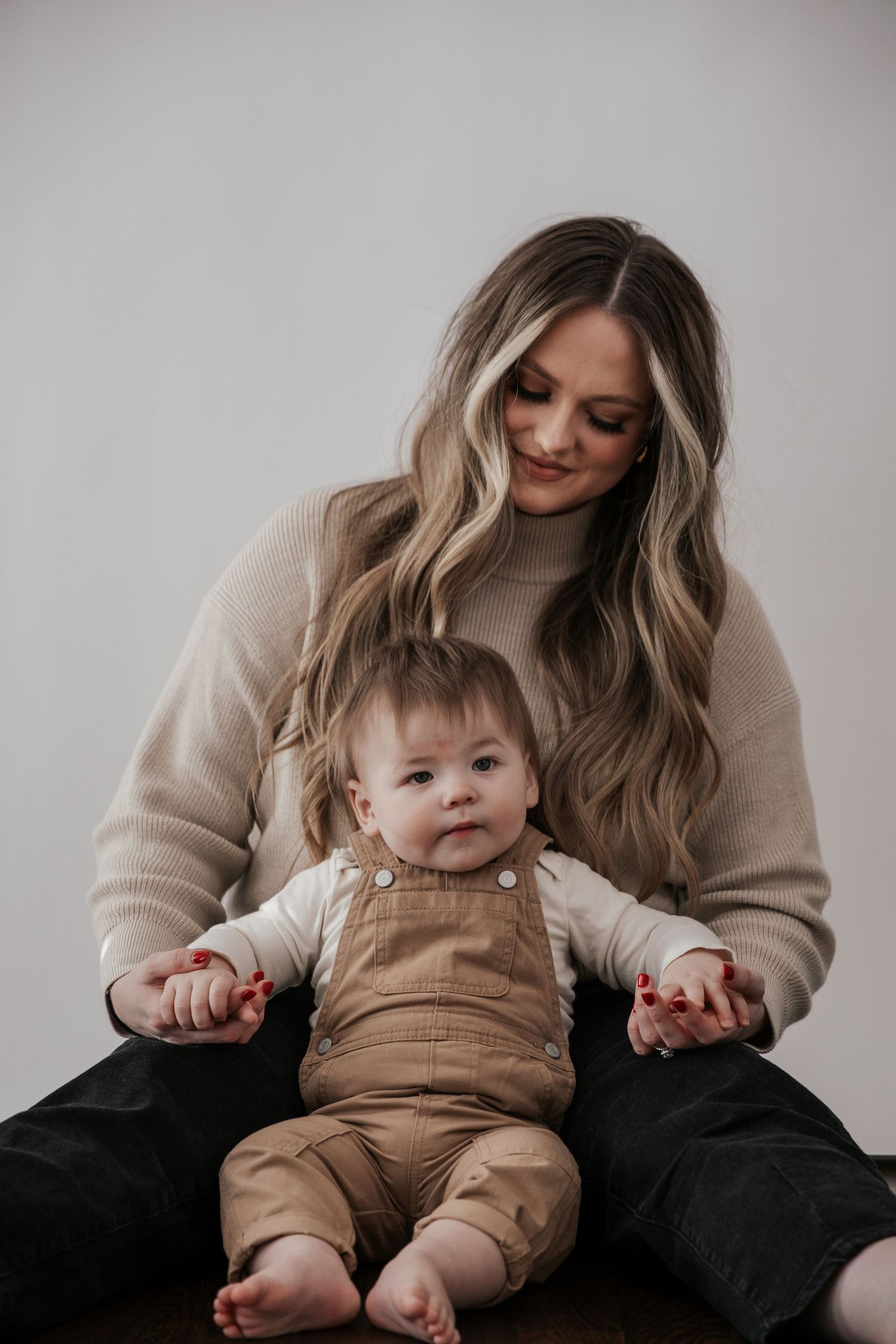 A smiling mother with long blonde hair holds her baby in brown overalls for a studio portrait.