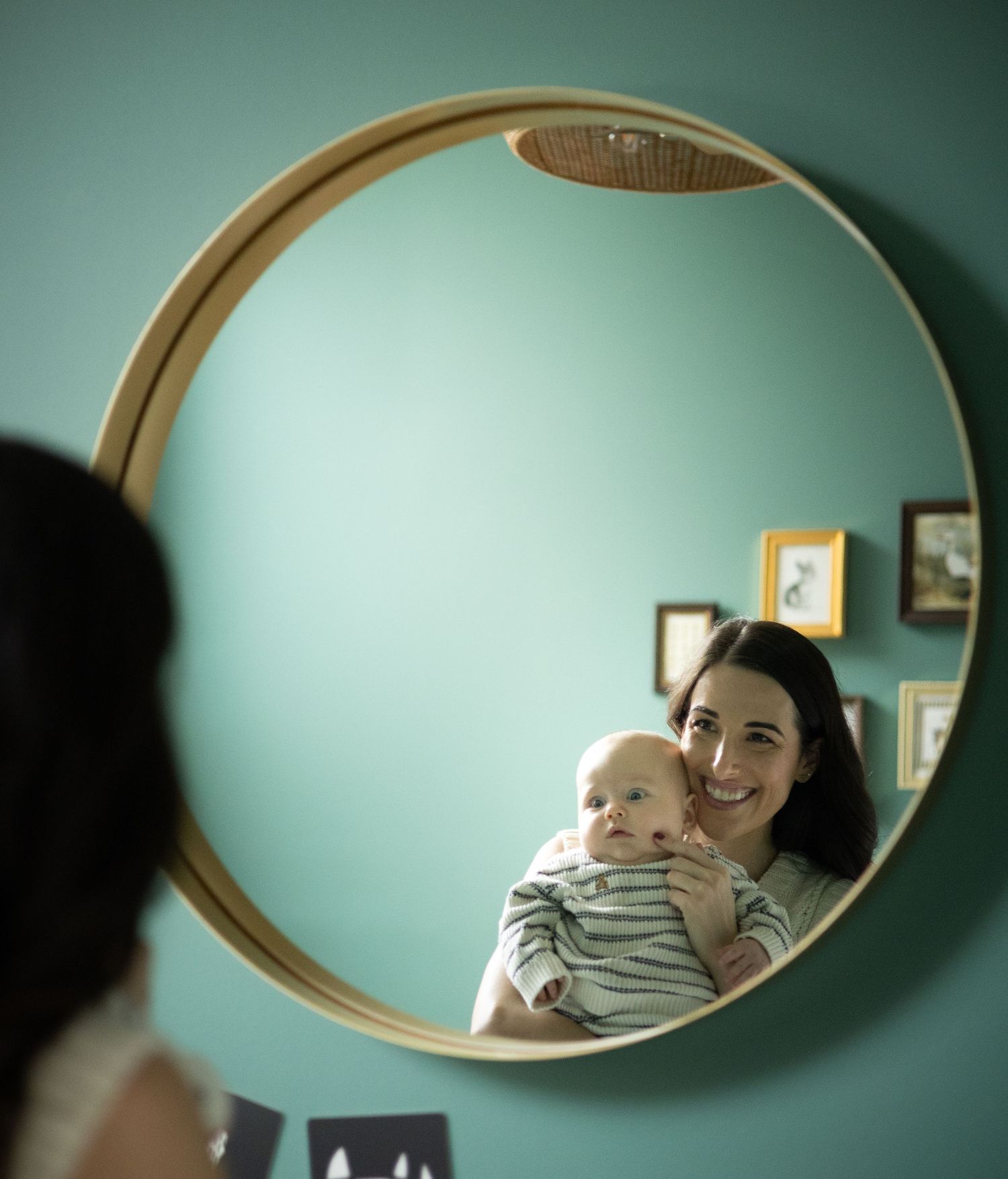 Joyful mother holding her smiling baby, both reflected beautifully in a round mirror on teal wall.
