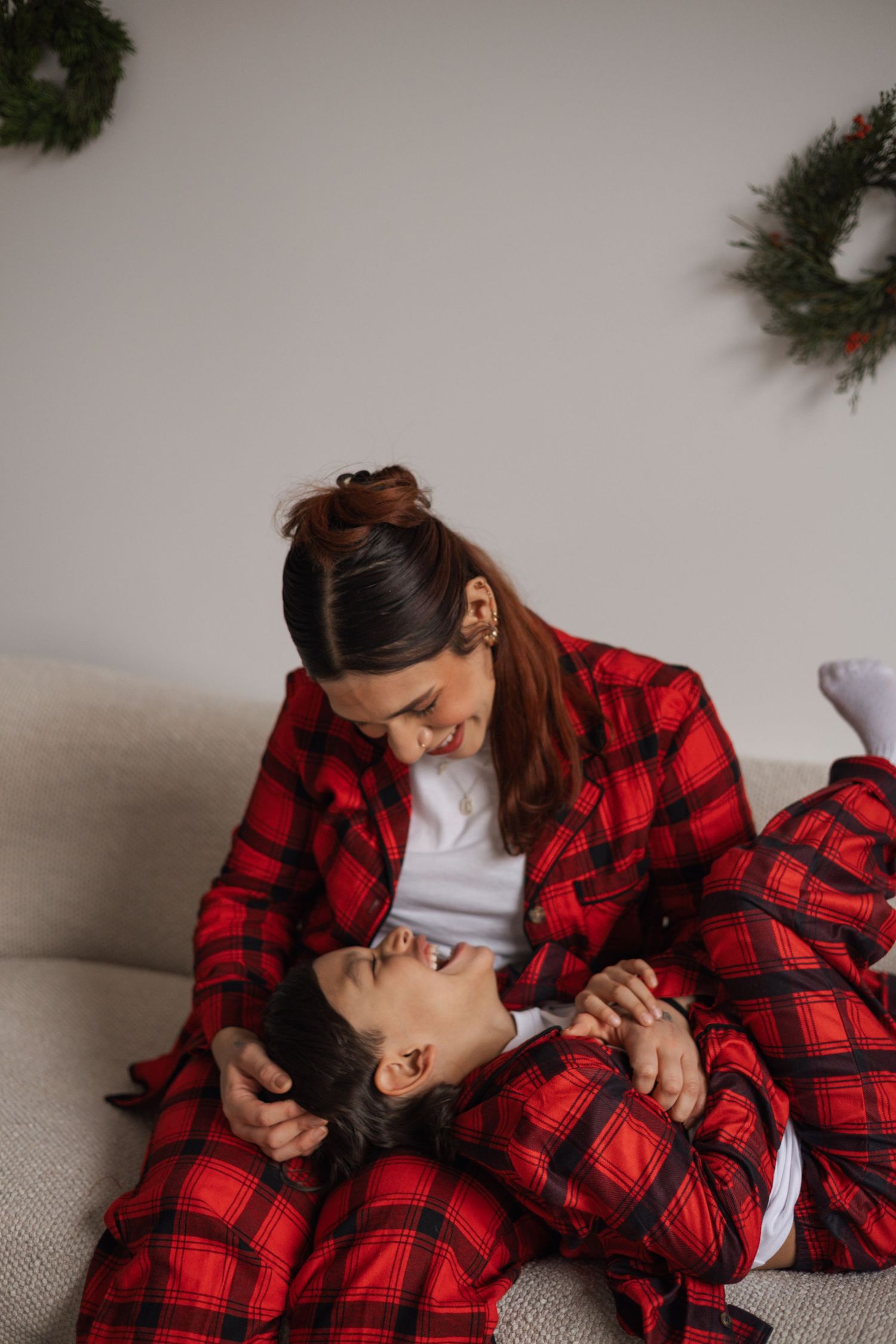 A mother and child in matching red plaid pajamas cuddle playfully on a cozy sofa at Christmas.