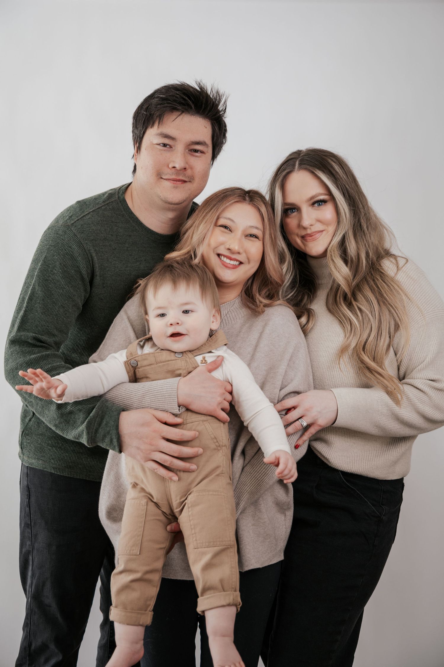 A family of four poses together in a bright studio, with two adults holding a baby in overalls.