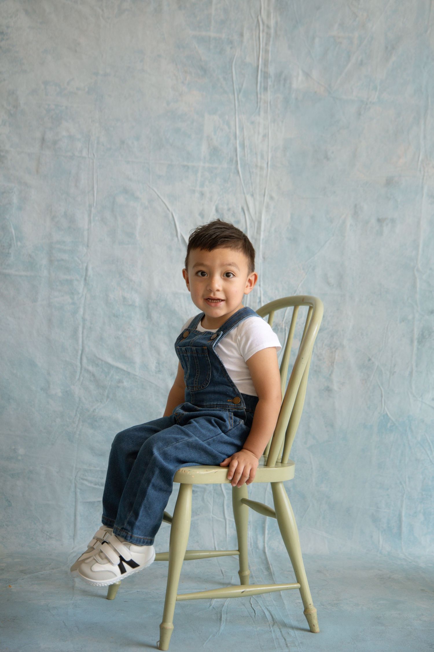 Young boy in denim overalls and white shirt sits on a green wooden chair against a blue textured backdrop.