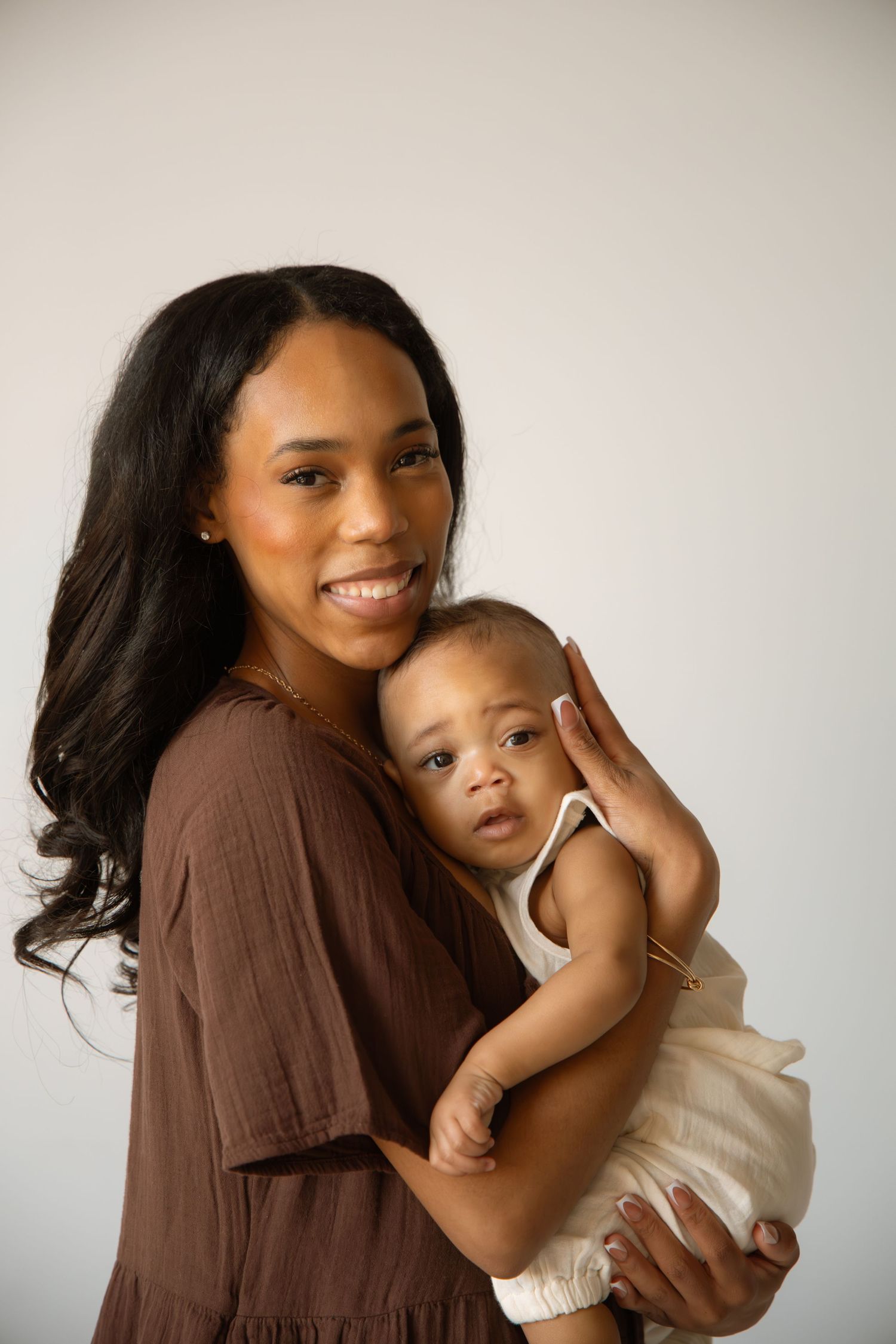 A smiling mother in a brown dress holds her baby in a white outfit against a light background.