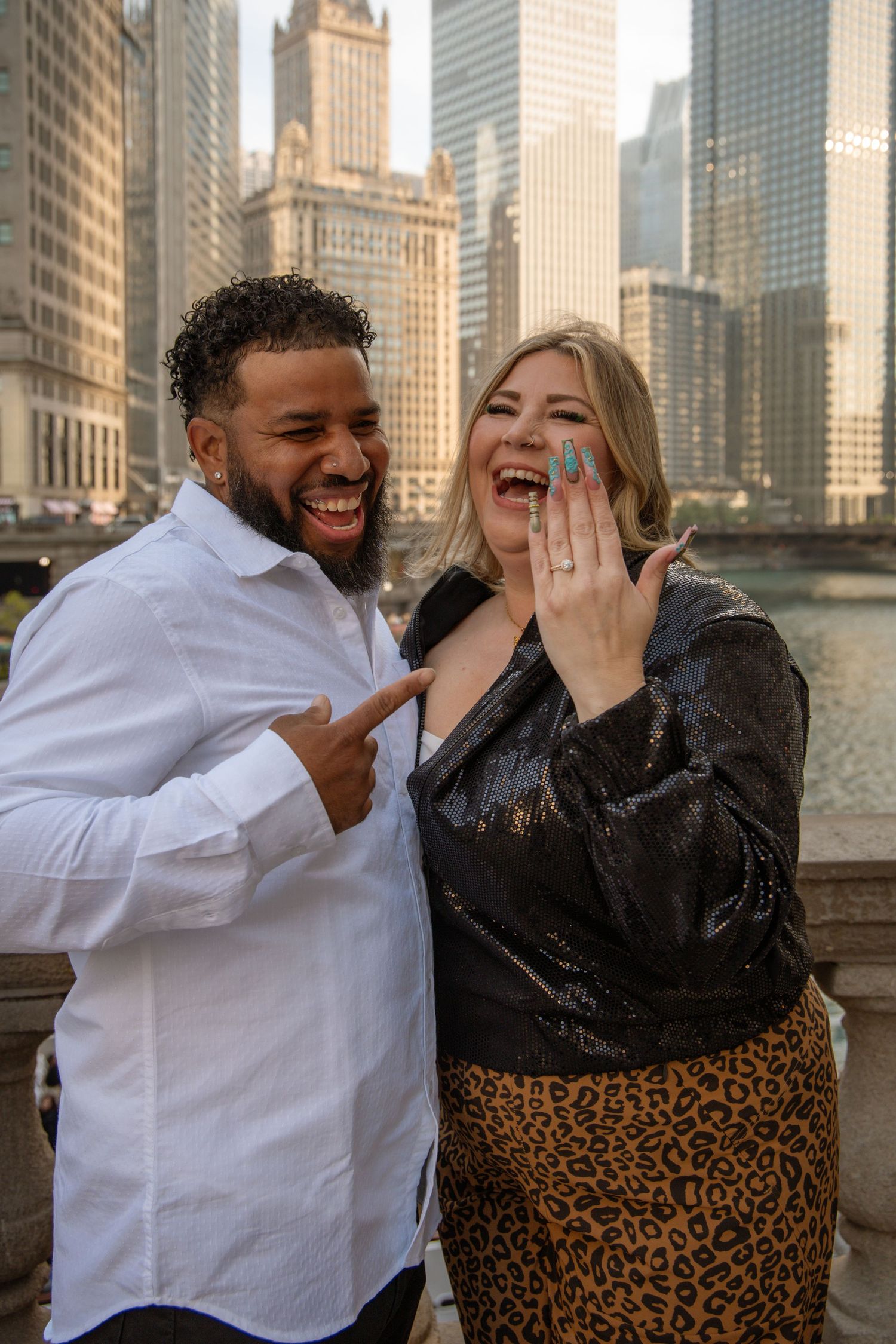 Joyful couple shows off engagement ring on Chicago Riverwalk, laughing together.
