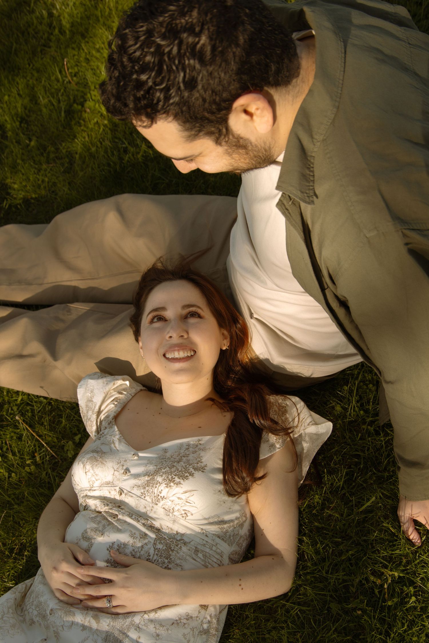 A smiling woman with red hair lies on grass while a dark-haired man in a tan suit gazes down at her tenderly.