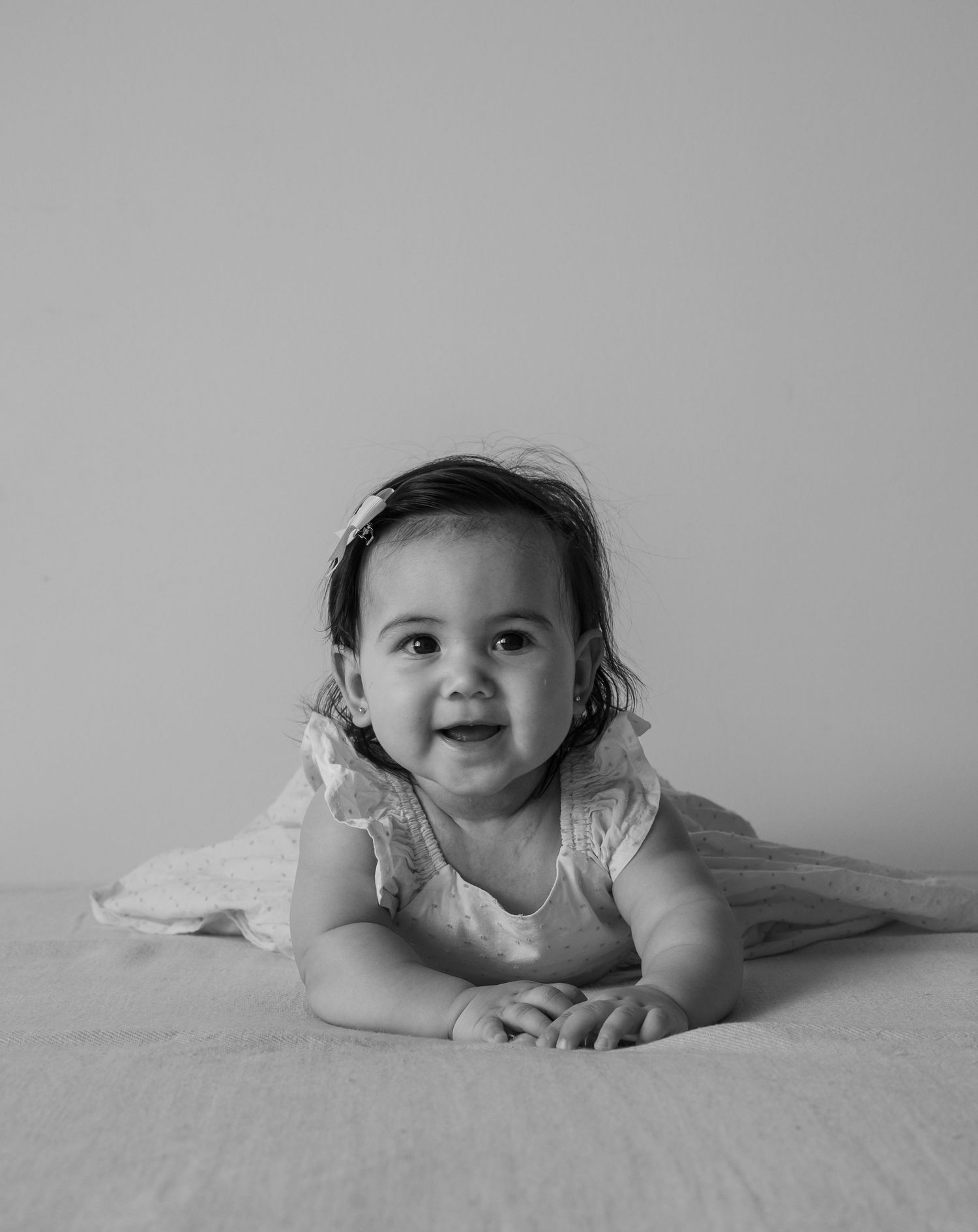 Black and white photo of a smiling baby girl in a lace dress doing tummy time on a white surface.
