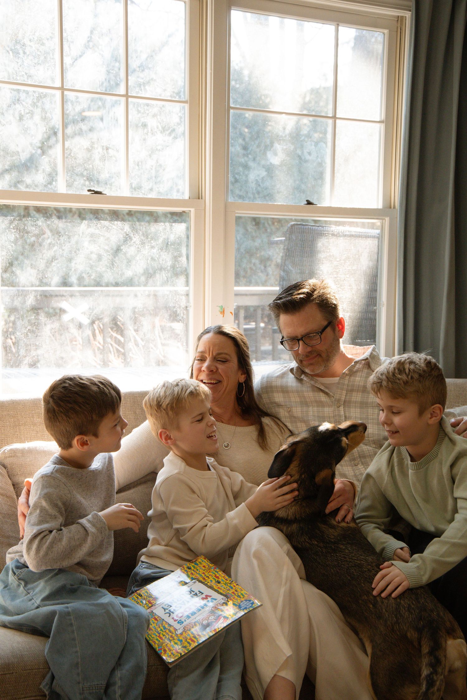 Family with three boys and a dog sitting together on a couch by a sunny window in a cozy living room.