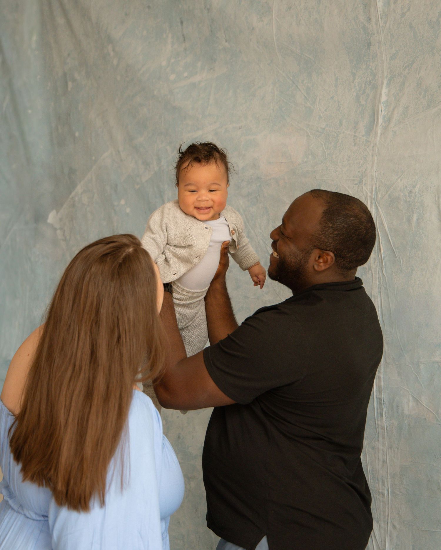 A joyful couple holds their smiling baby up in the air against a soft blue-gray backdrop.