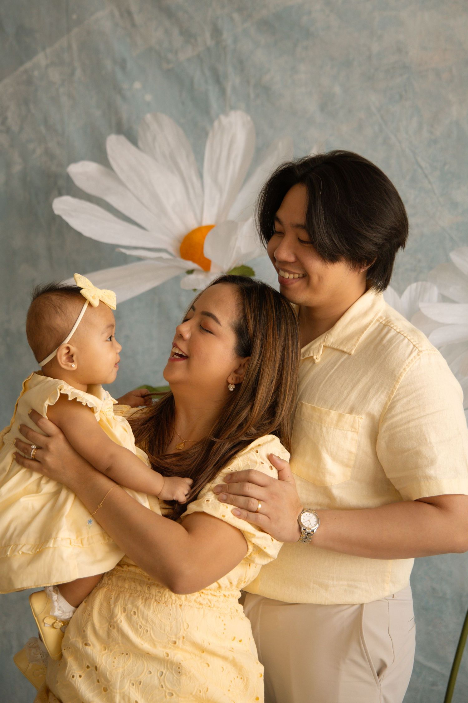 Happy Asian family in yellow outfits posing with baby girl before floral backdrop.